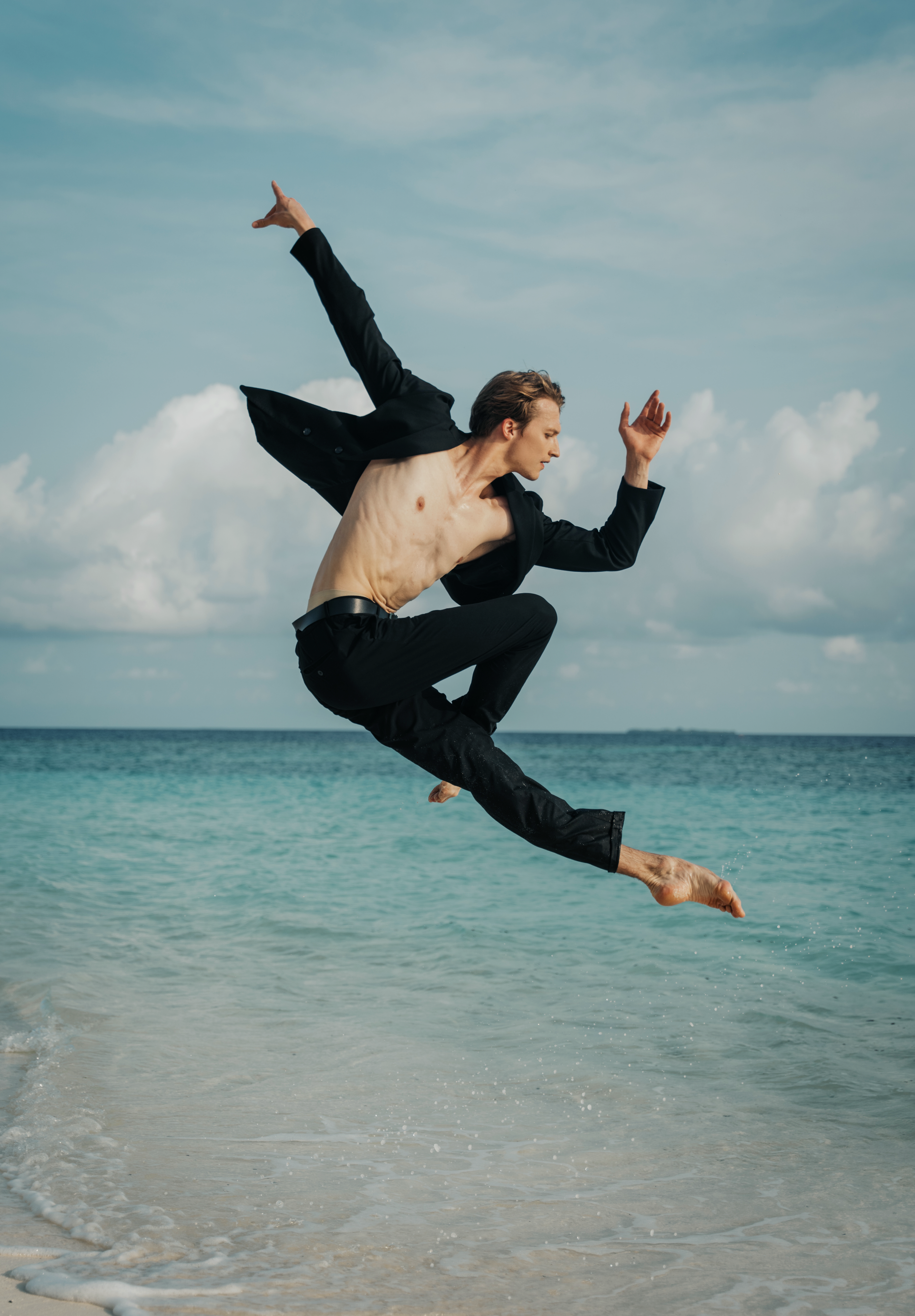 Man in suit jacket and pants dances barefoot on beach, arms outstretched, blue sky and ocean background.
