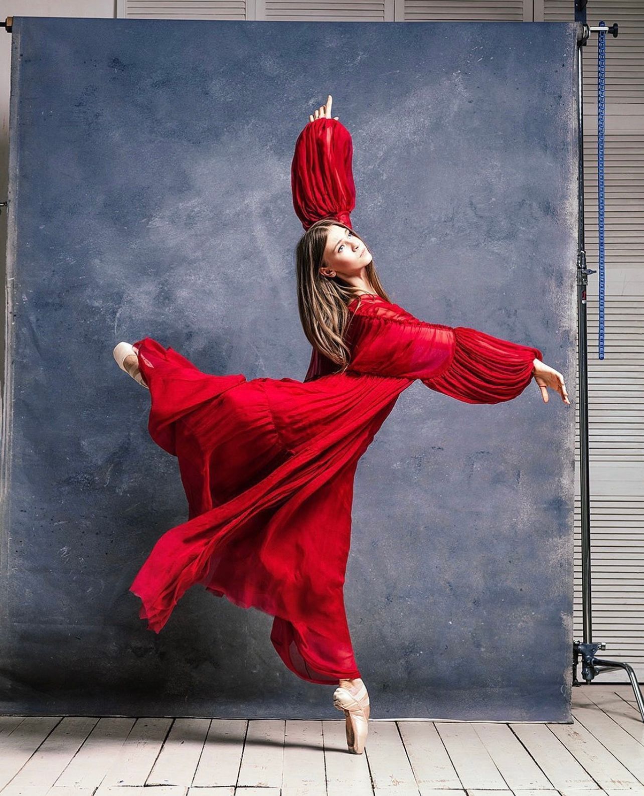 Ballerina in red dress, en pointe, arms extended, against a blue backdrop.