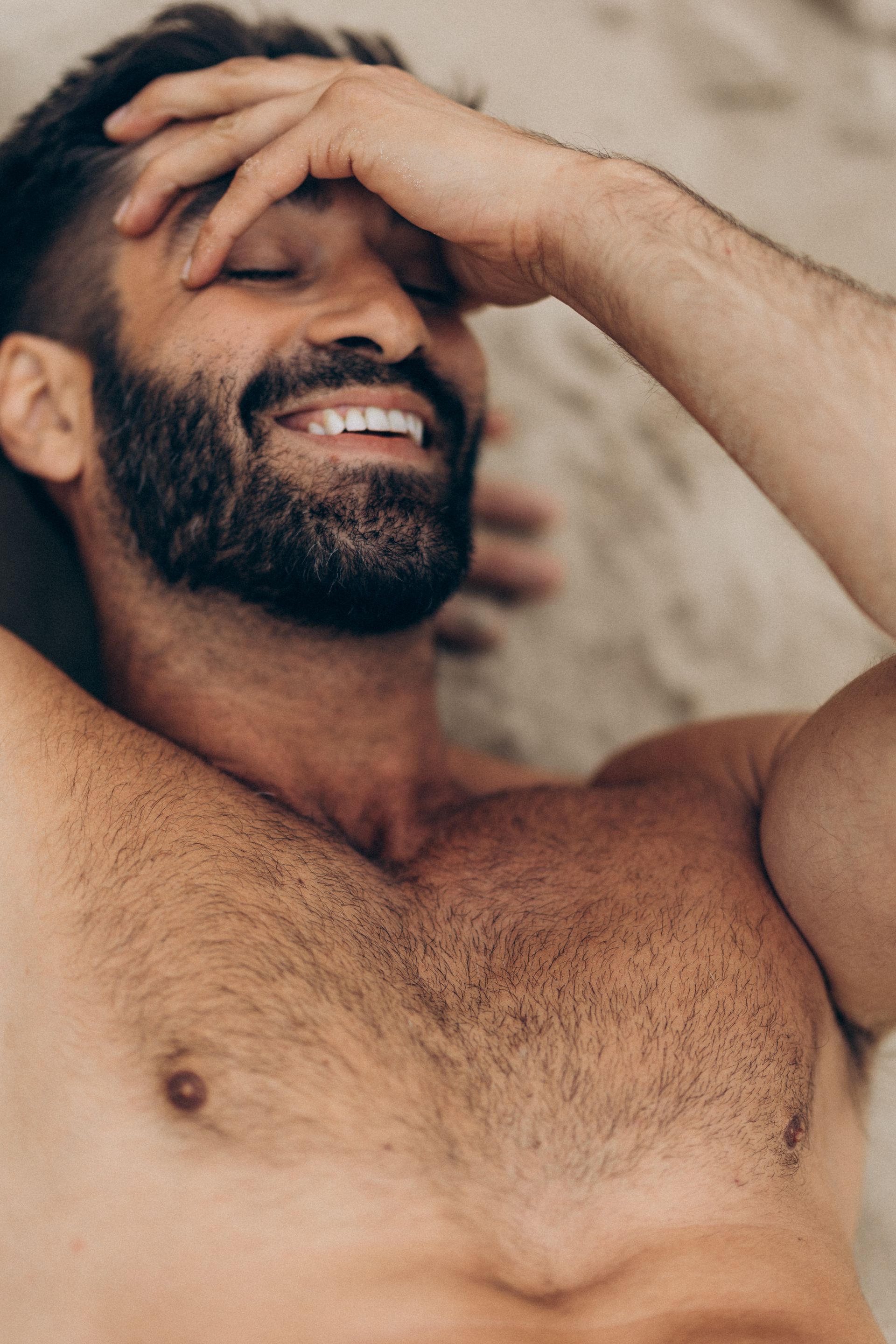 Man with a dark beard smiles, hand on forehead, lying shirtless on sand.
