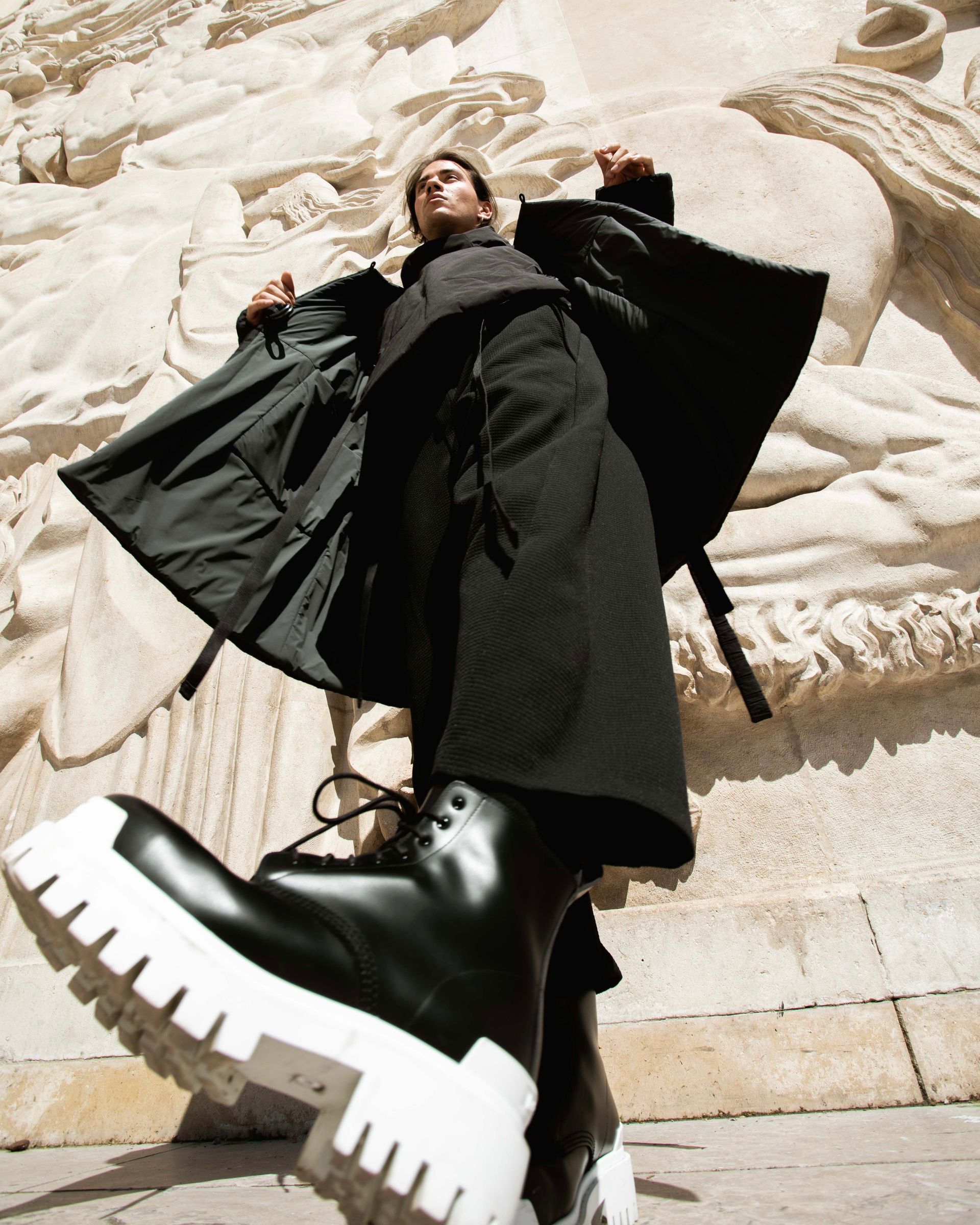 Person in black coat and massive white-soled boots poses dramatically against a stone relief.