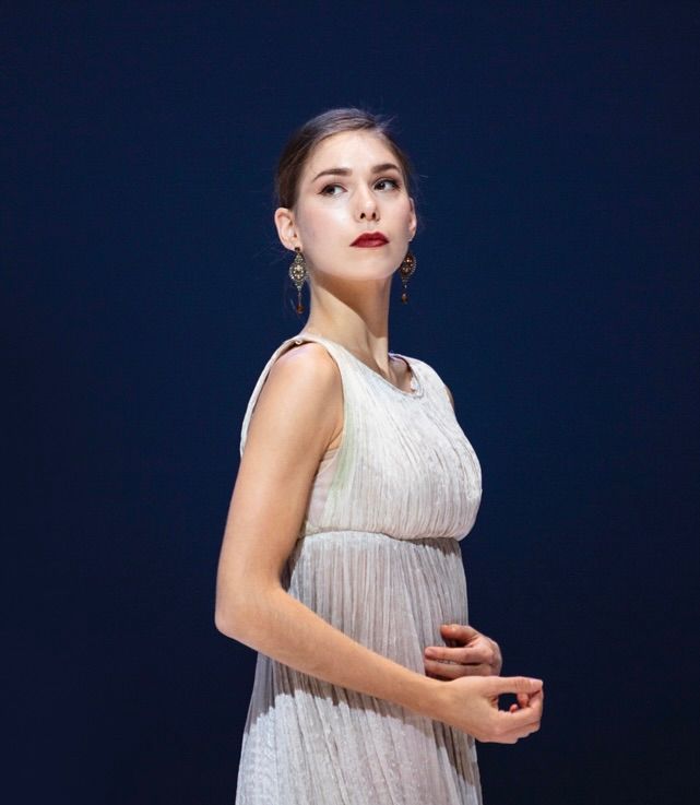 Woman in white dress, looking upward, hands clasped. Dark background, wearing gold earrings and red lipstick.