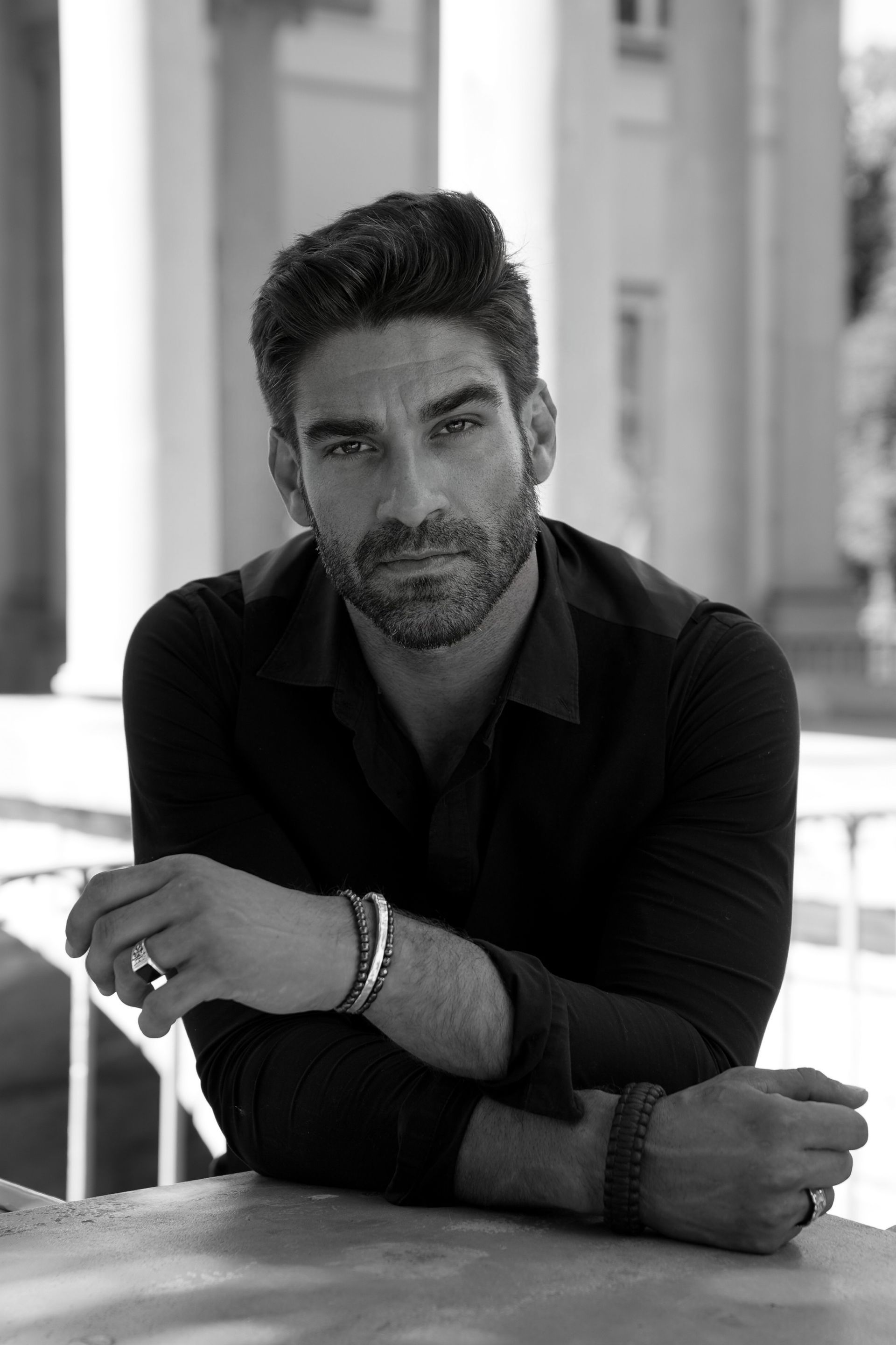 Man with dark hair and beard, arms crossed on a table, looking at the camera. Black and white photo outdoors.