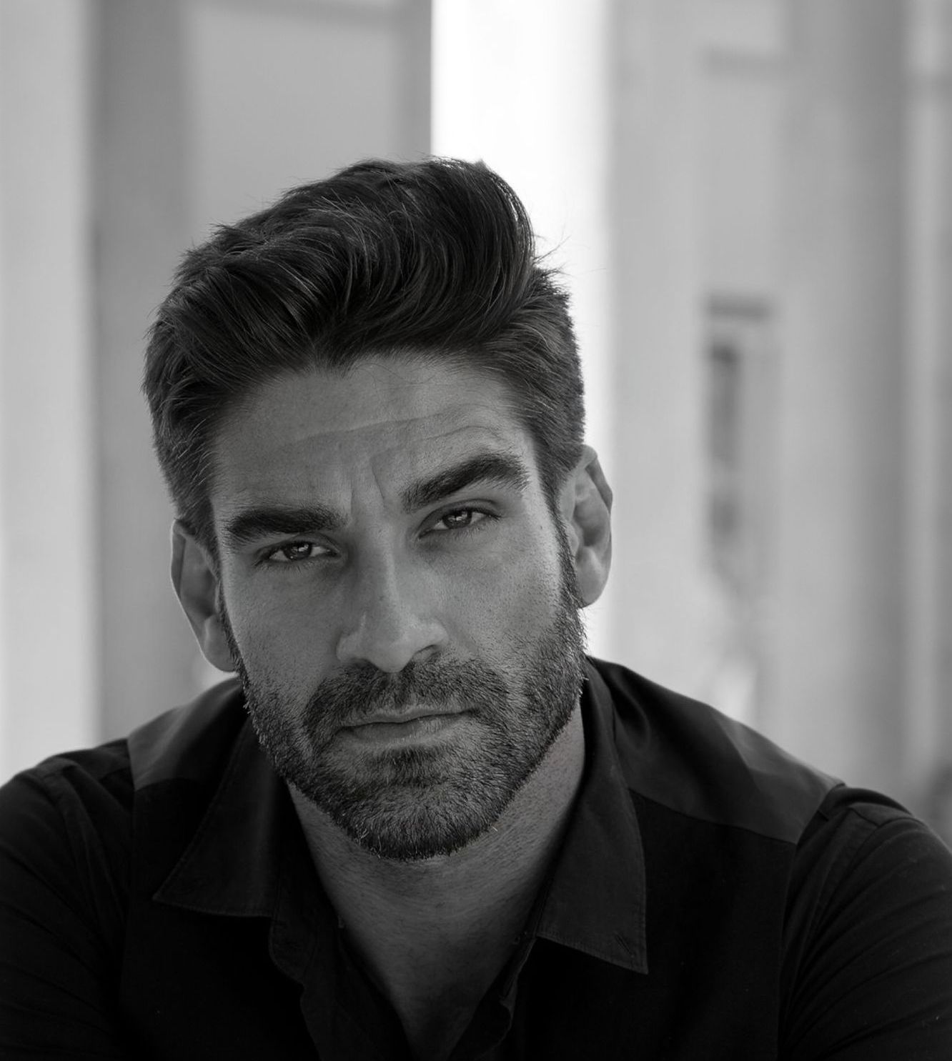 Man with dark hair and beard, wearing a black shirt, looking at the camera. Black and white photo.