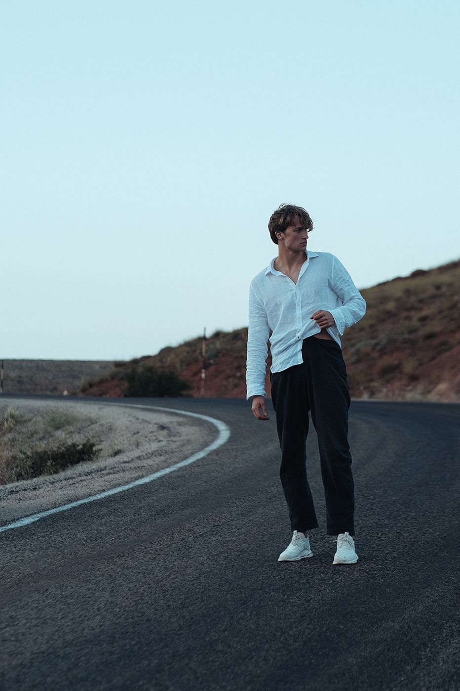 Person stands on a curved road, wearing a white shirt and black pants, looking off to the side.
