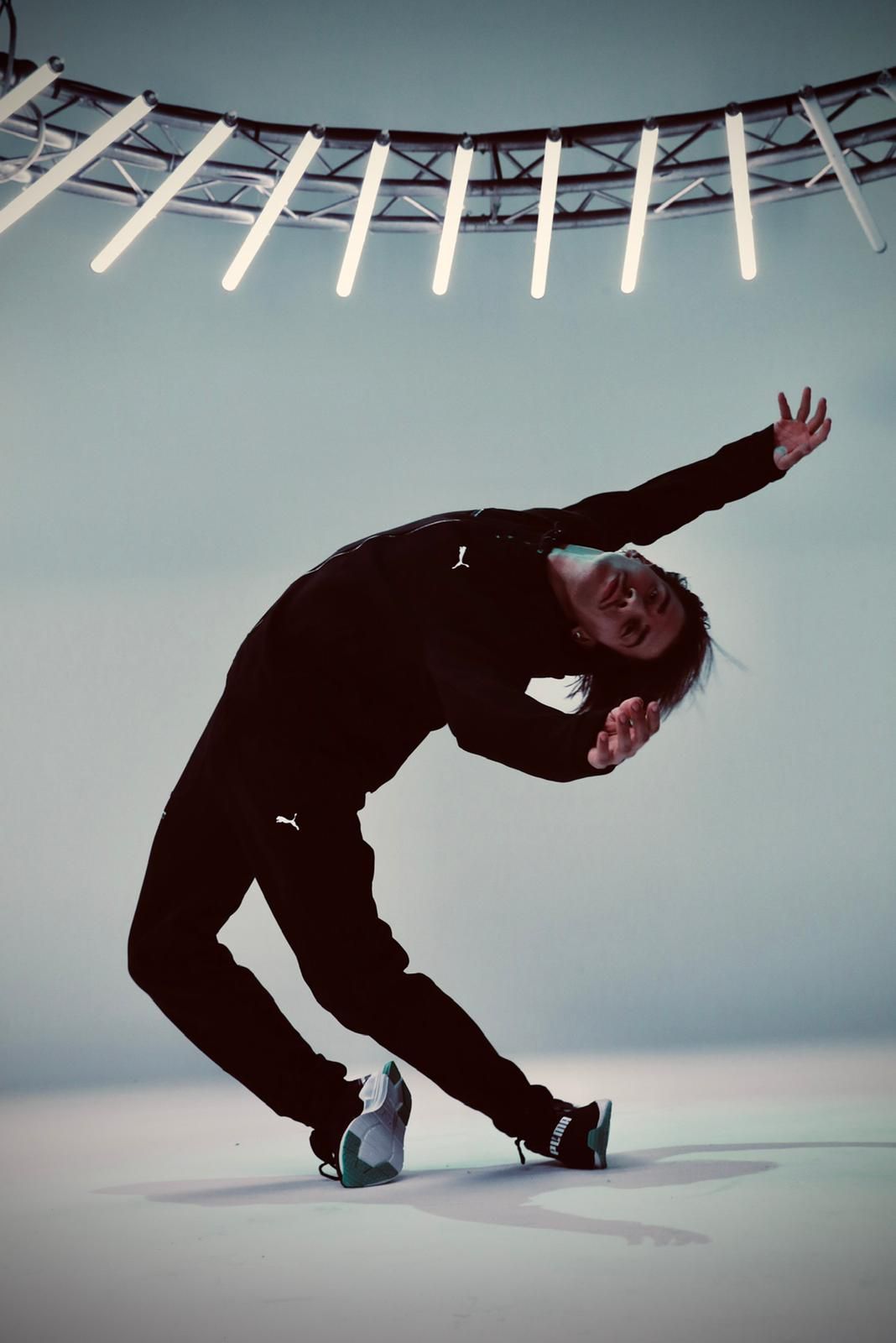 Dancer in black outfit, arched backward, arms outstretched. Lit stage background.