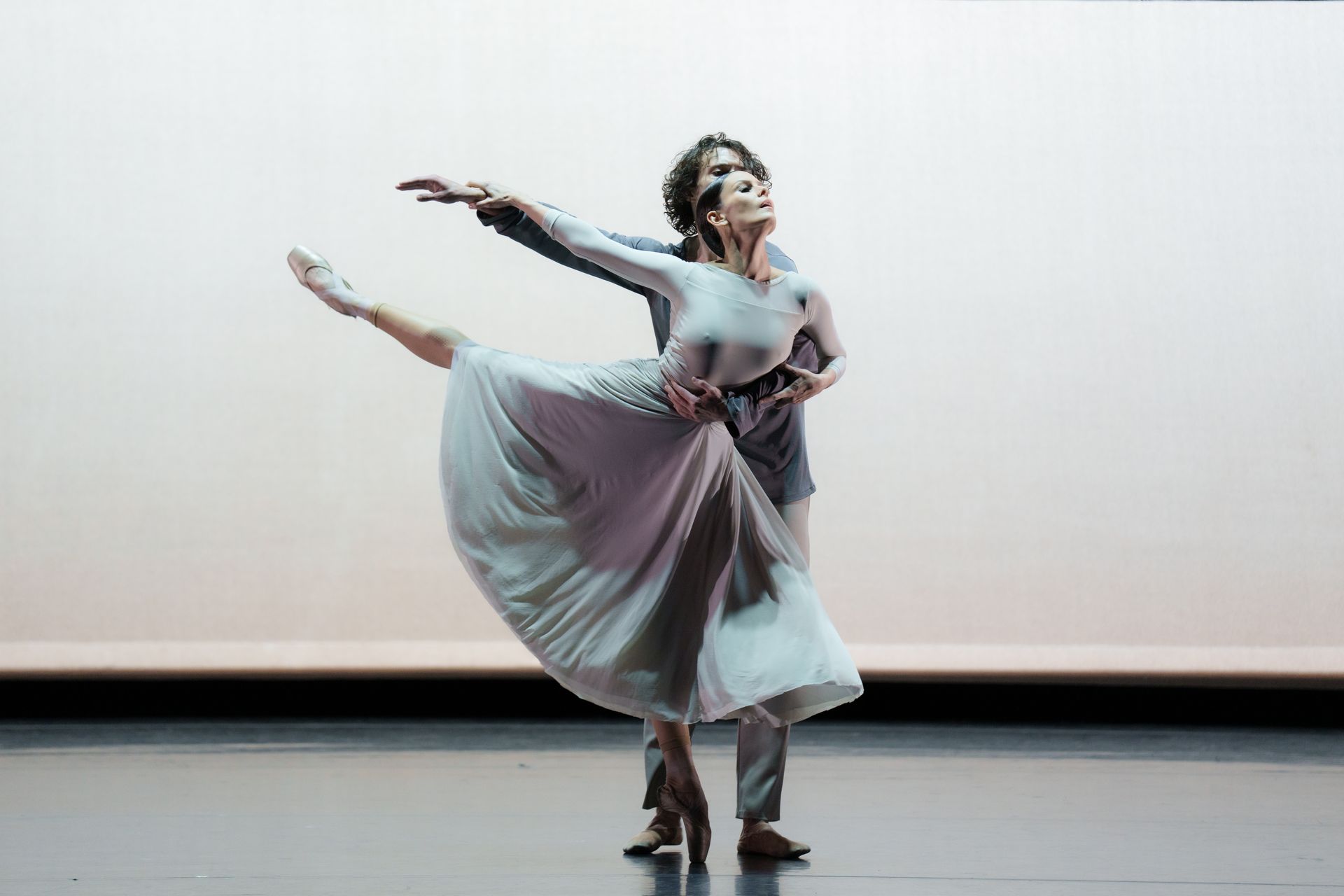 Ballet dancers performing on stage: woman in white dress, man in suit, arms outstretched, neutral background.