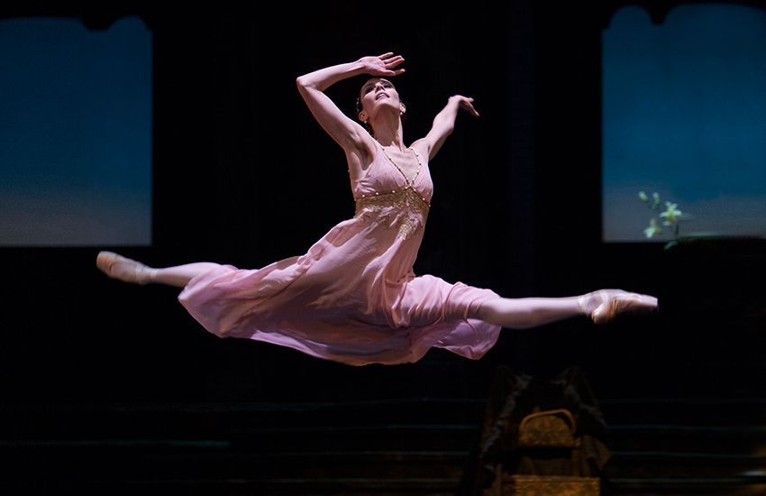Ballerina in mid-air split, wearing a pink dress, arms outstretched, performing on stage.