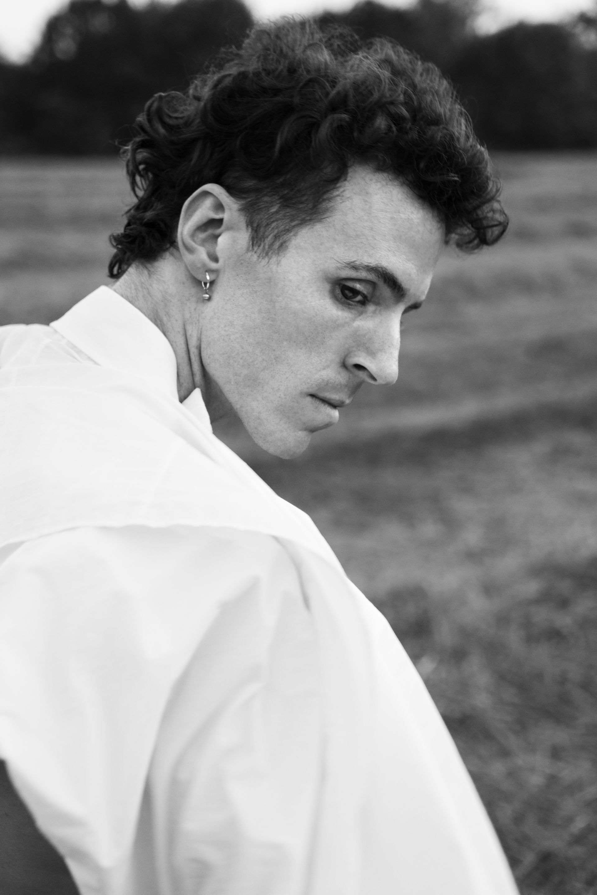 Man in white shirt looking down, outdoors. Curly hair, earring. Black and white.