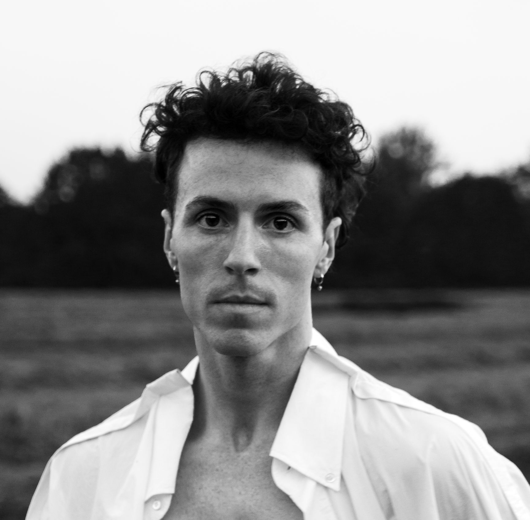 Man with curly hair, open white shirt, field backdrop.