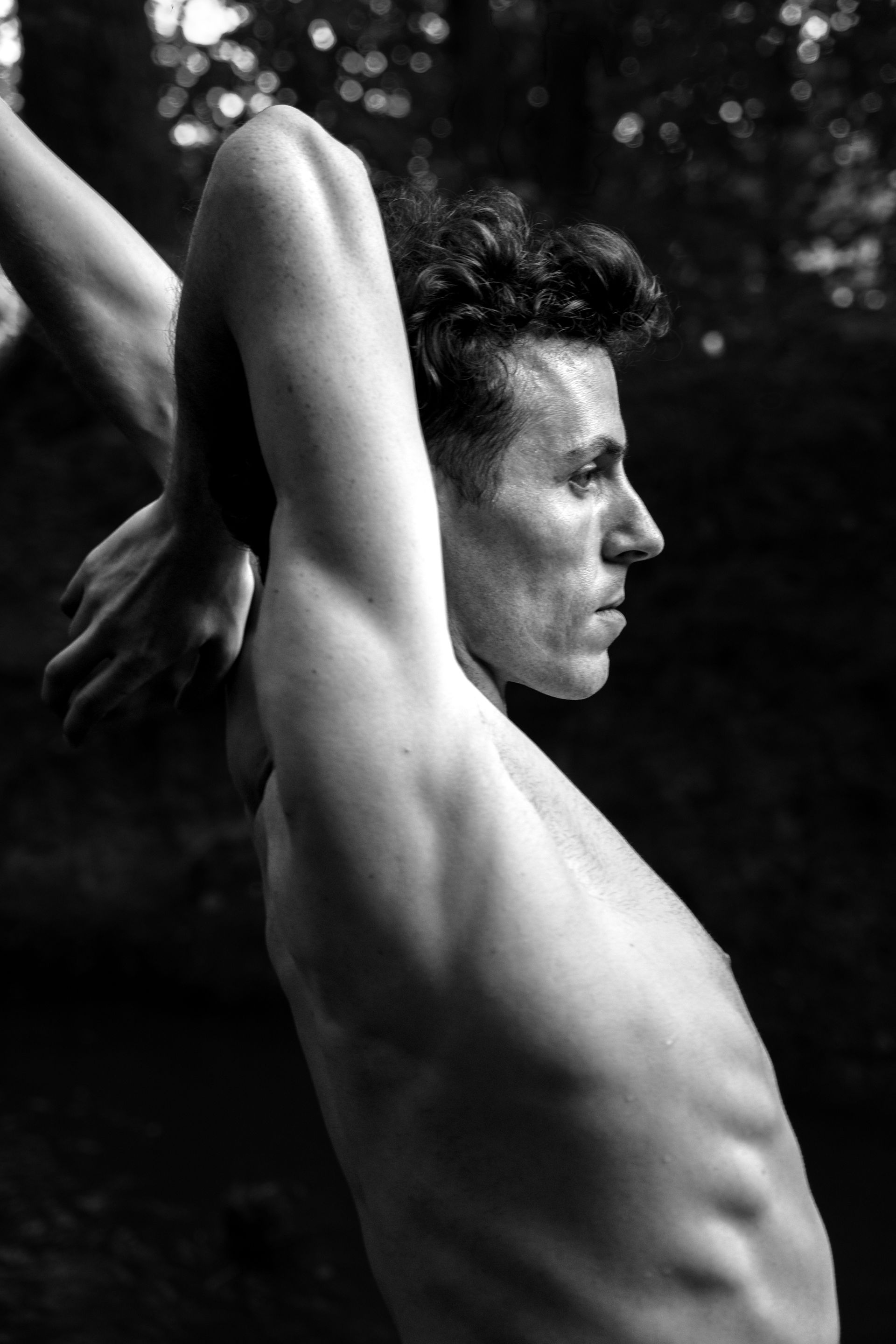 Man arches back, arms overhead, profile view. Outdoors, shirtless. Black and white photo.