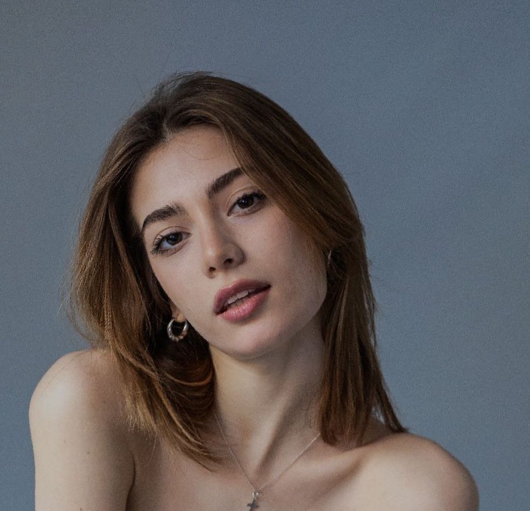 Woman with light brown hair, looking at the camera. Wearing a necklace and hoop earrings, with a neutral background.