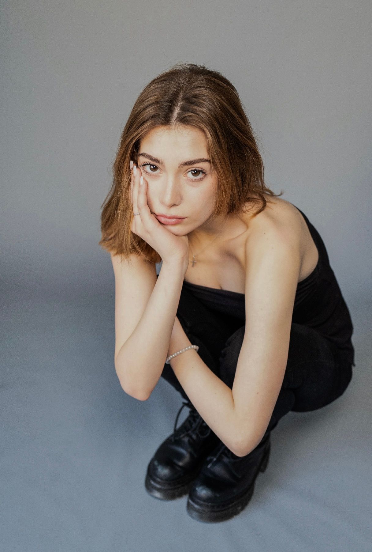 Young person in black outfit and boots, hand on face, looking at camera against gray background.