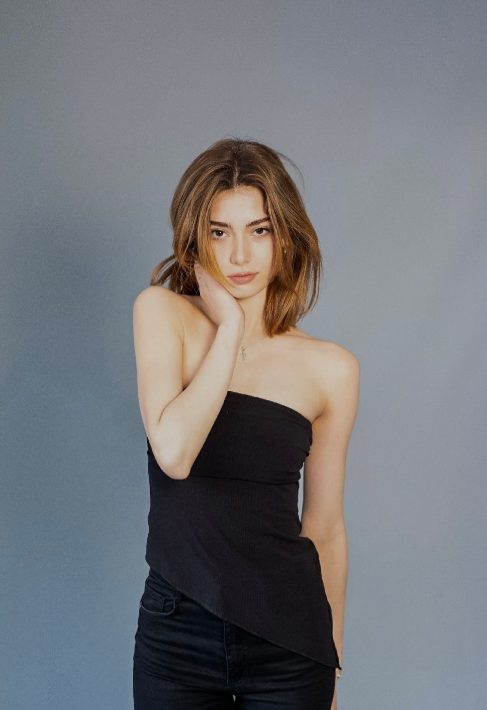 Woman in black strapless top, holding her cheek, against a gray background.