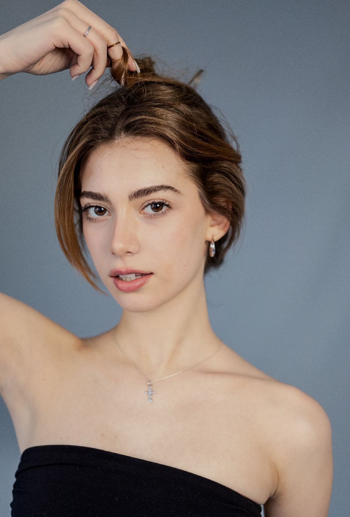 Woman with fair skin, pulling up her hair; wearing a strapless black top and silver jewelry; gray background.