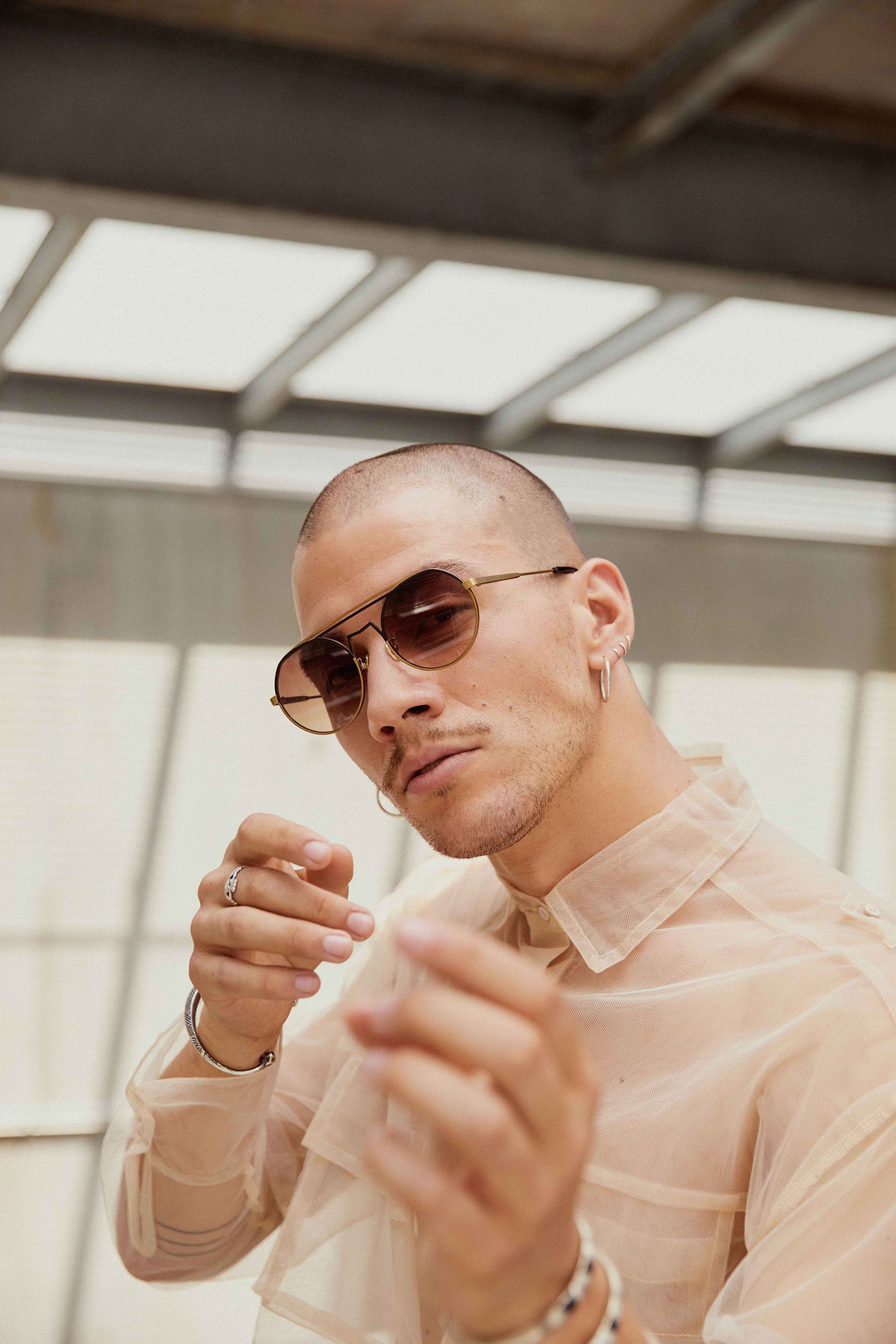 Man in sheer beige shirt and sunglasses, looking towards the camera, hands raised.