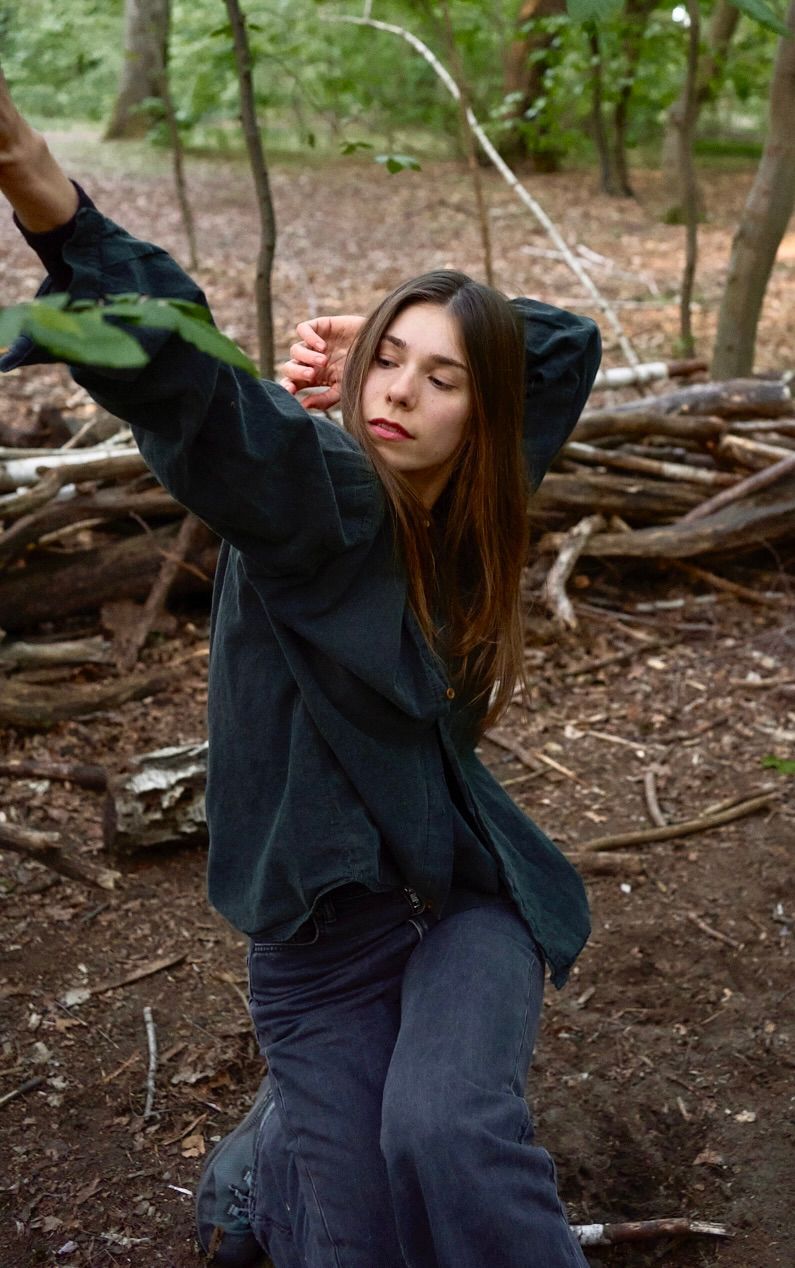 Woman in denim shirt and jeans, reaching for leaves in a forest, eyes closed.
