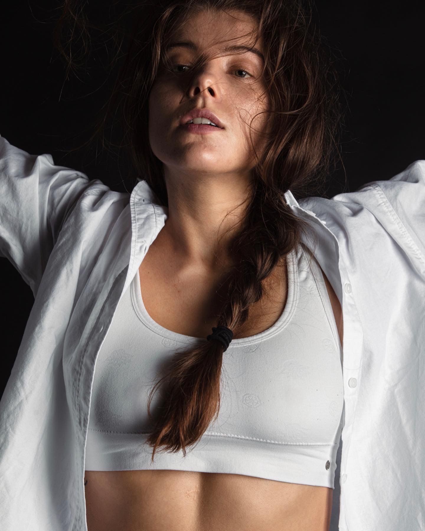 Woman in white bra and open shirt, arms raised, dark background.