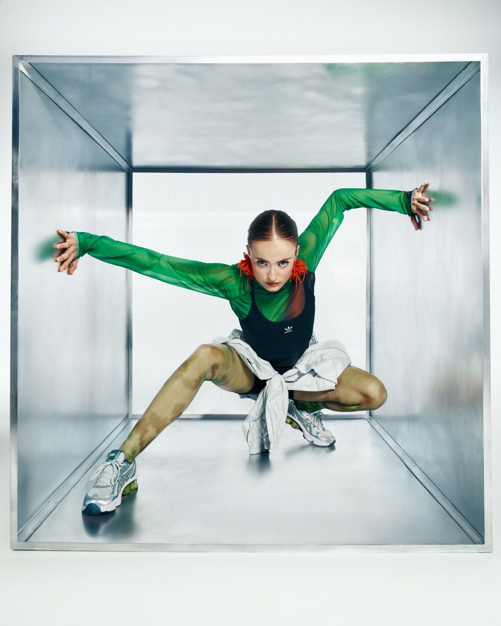 Woman in athletic wear poses inside a metal cube with arms outstretched, green shirt, and sneakers.