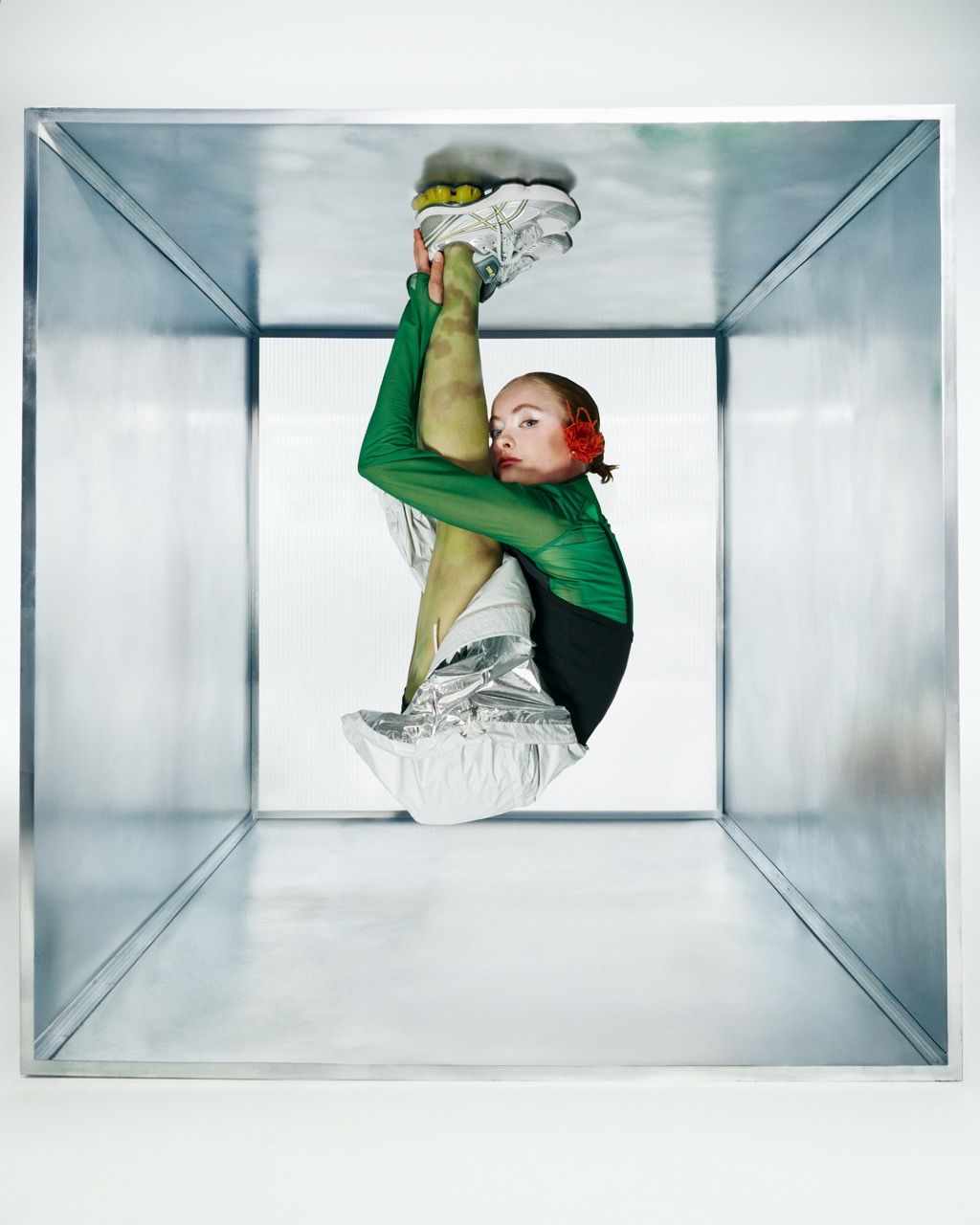 Person inside a metallic cube, legs extended. Wearing a green top, silver boots, and red ear adornments.