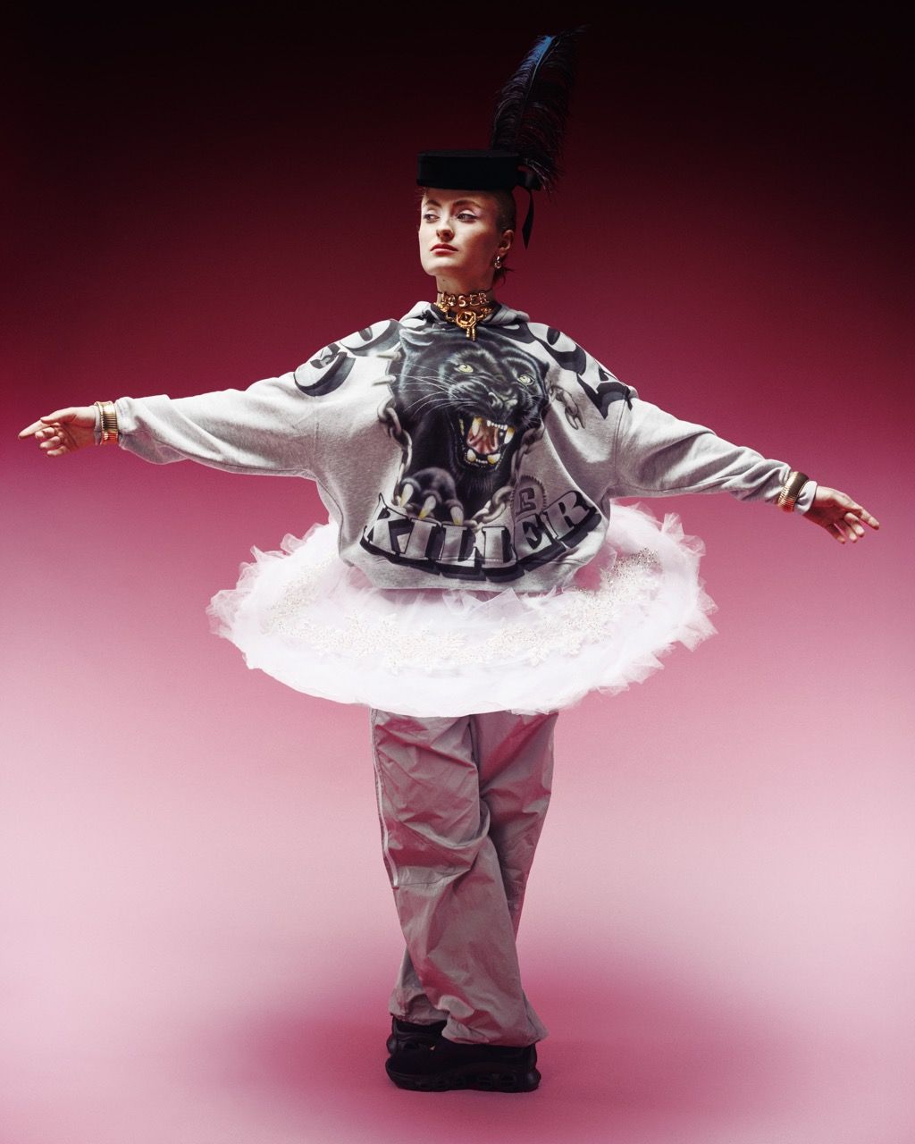 Person in sweatshirt, tutu, and pants strikes a ballet pose against a pink backdrop.
