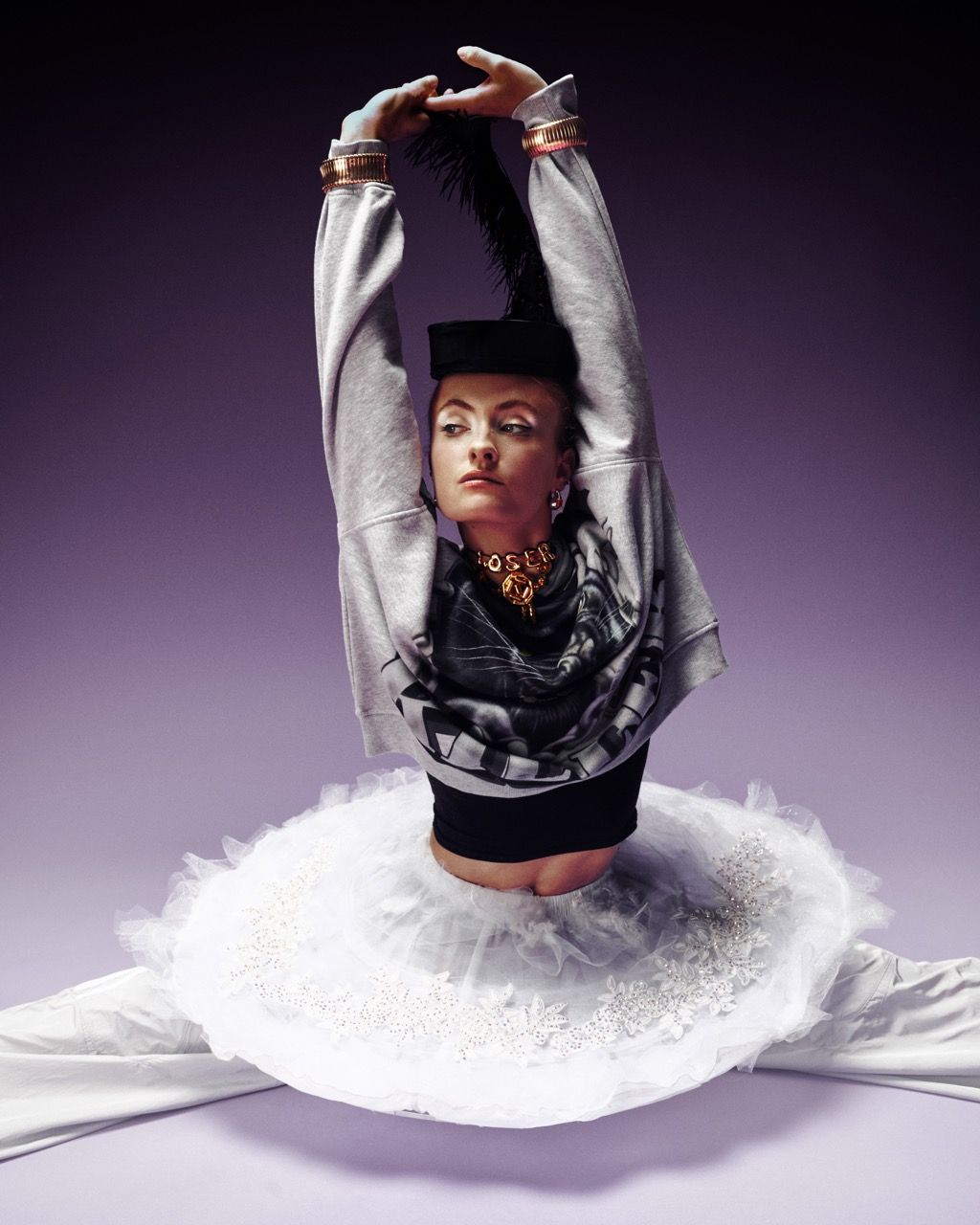 Bust of person in a tutu and sweater, arms raised, against a purple background.