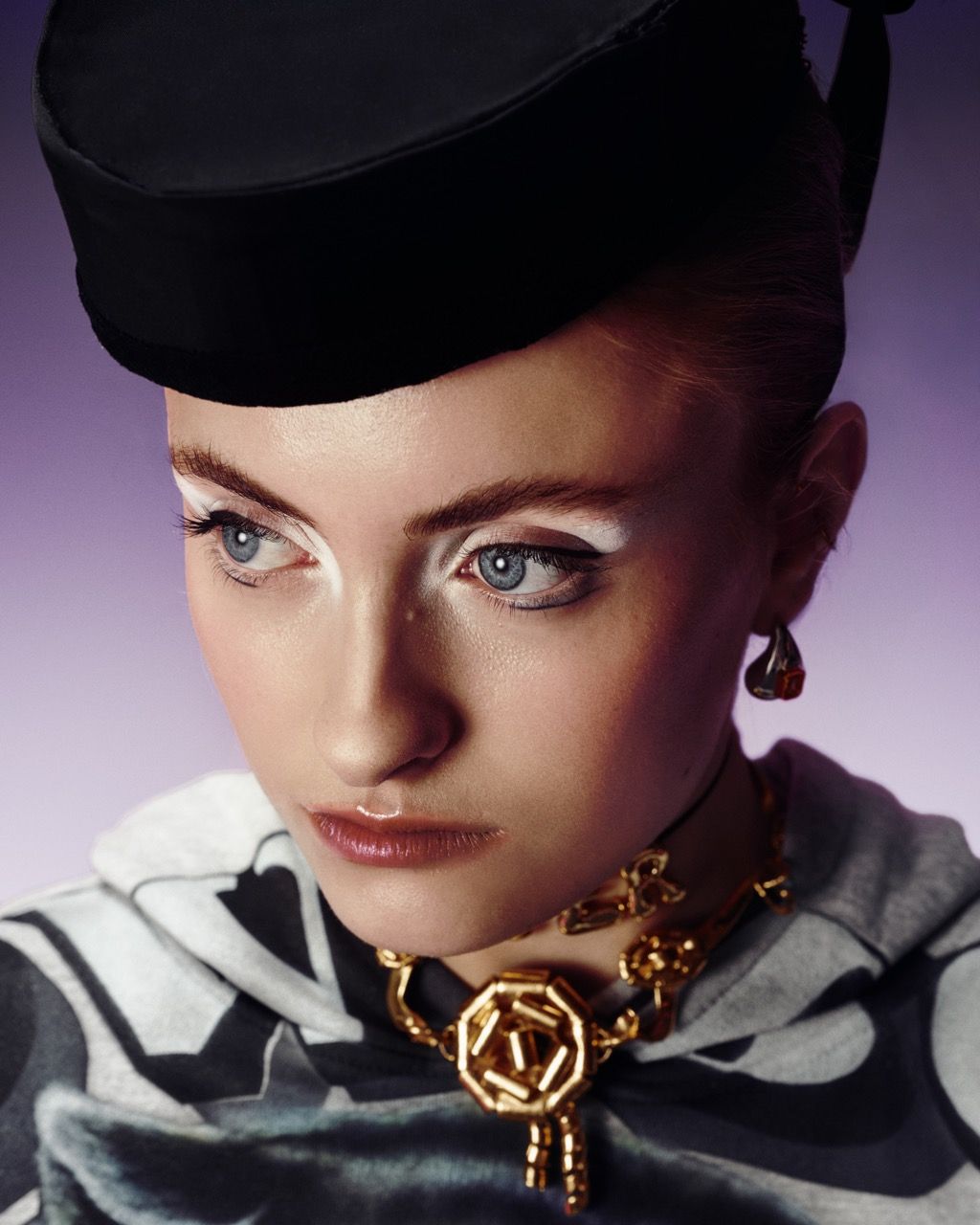 Woman wearing a black hat, gold jewelry, and white eye makeup, looking away, against a purple backdrop.