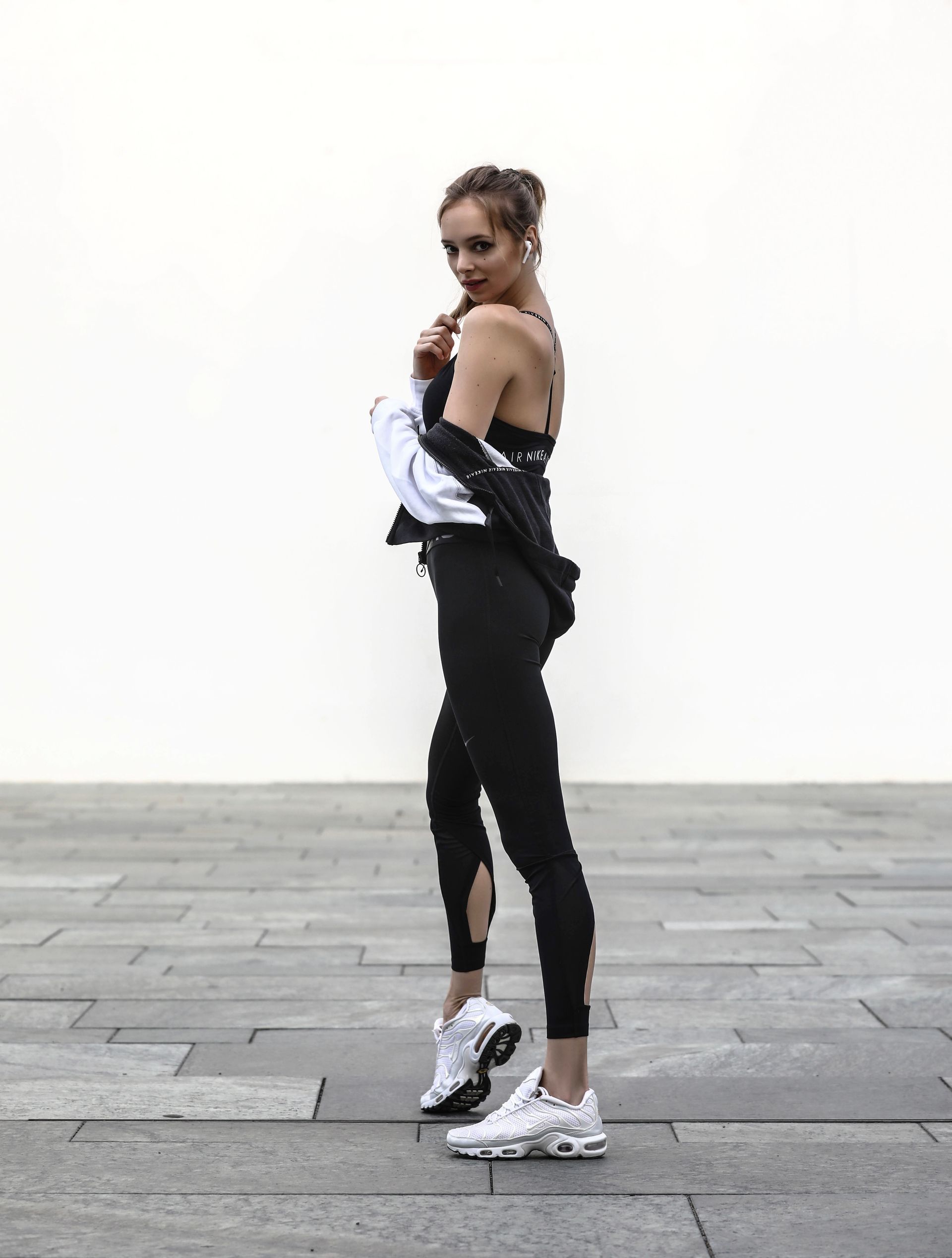 Woman in black activewear and sneakers poses outdoors, looking over shoulder.