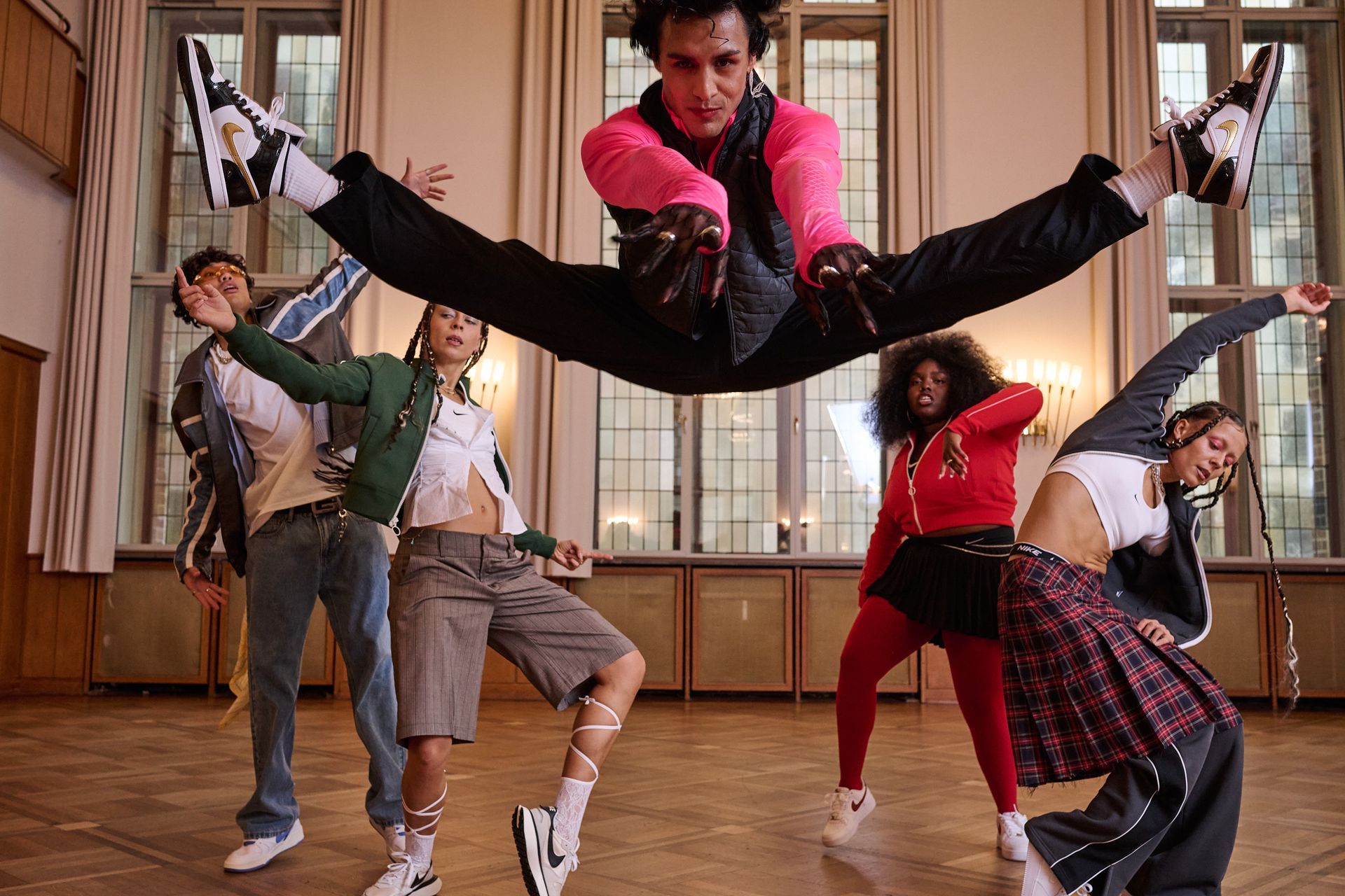 Five dancers in athletic wear pose dynamically in a room with large windows; one performs a split in mid-air.