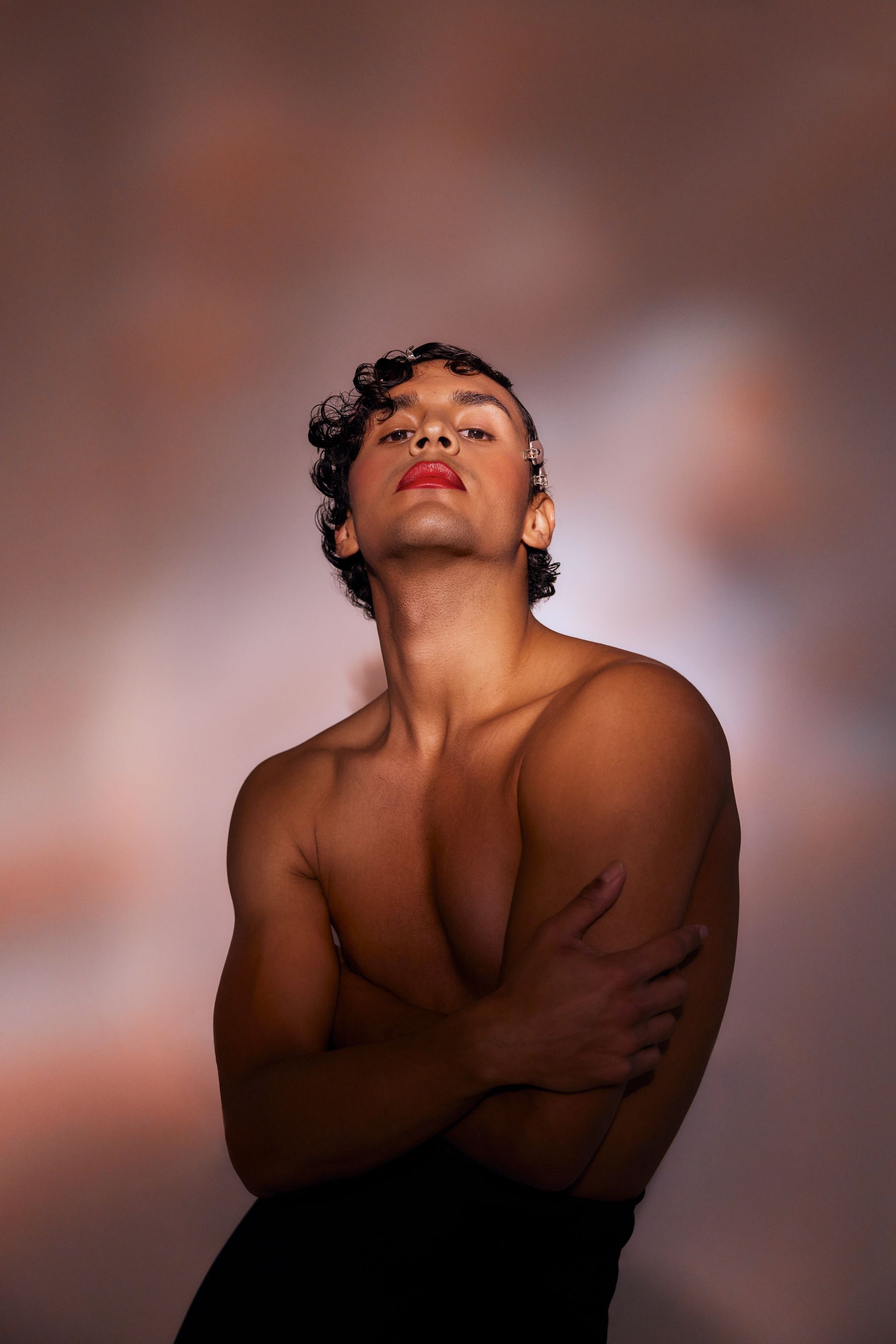Man with dark curly hair and red lipstick, arms crossed. Soft, neutral background.
