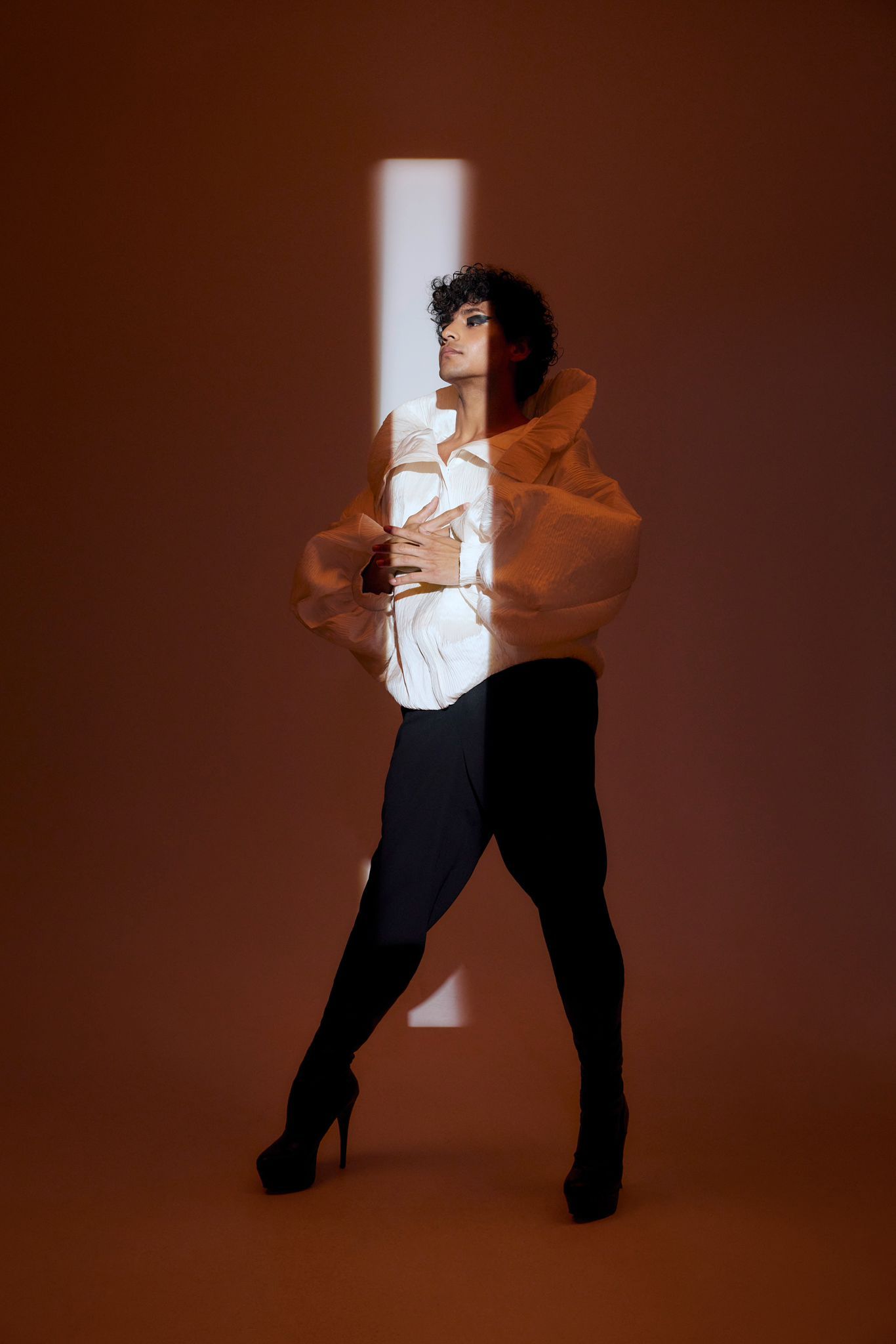 Person in black pants and high heels, wearing a white top and orange structured jacket, standing with an artistic backdrop.