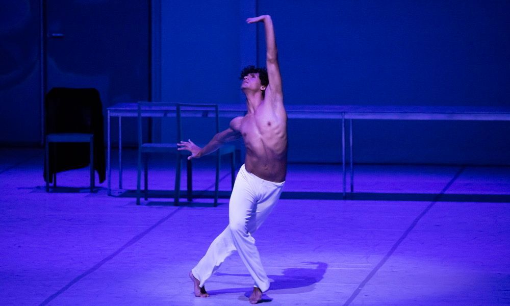 Male ballet dancer in white pants, shirtless, performing on stage with blue lighting.