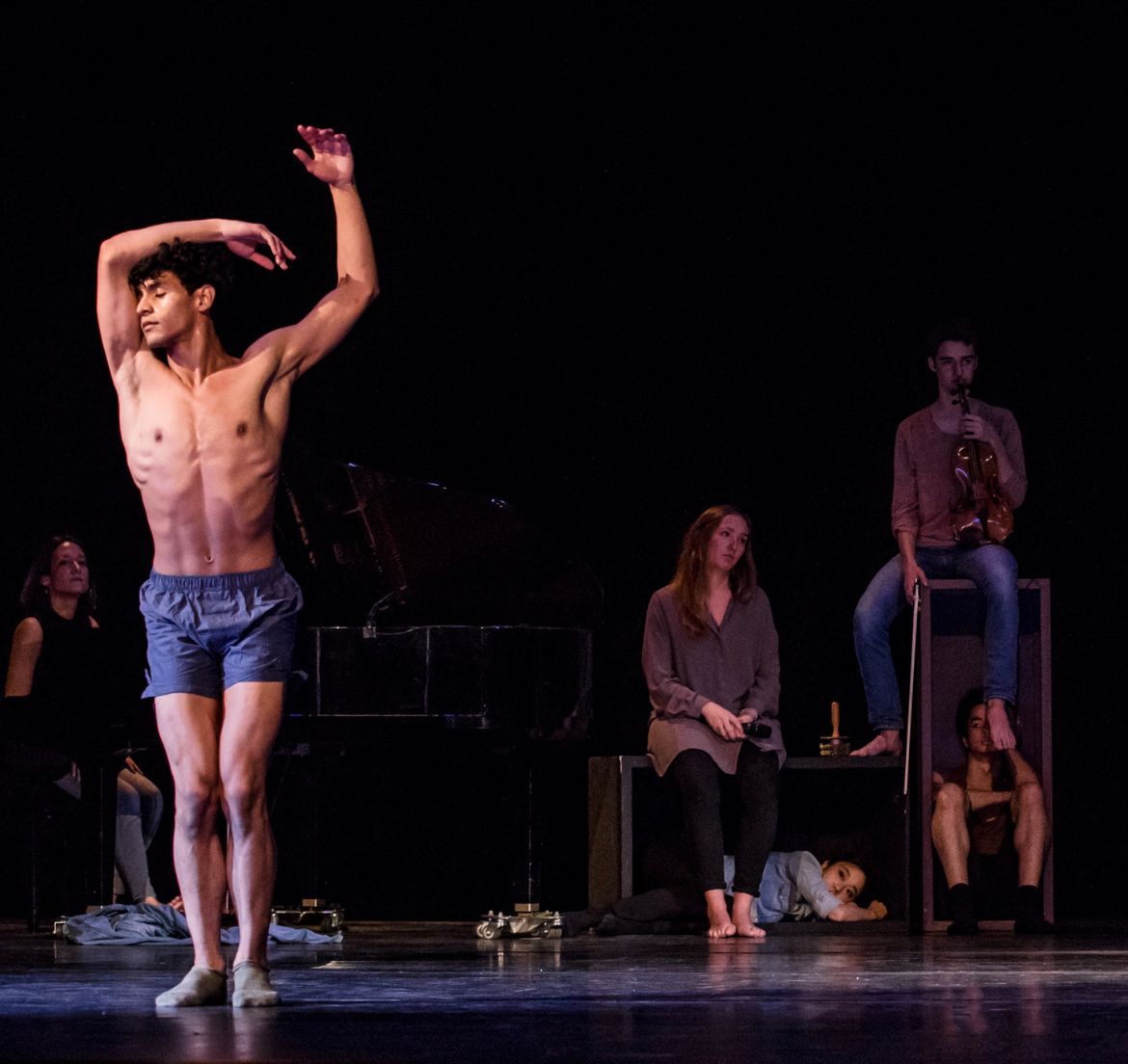 Dancer with raised arm, performing on stage. Other figures sit, stand, and recline near a piano, musician, and objects.
