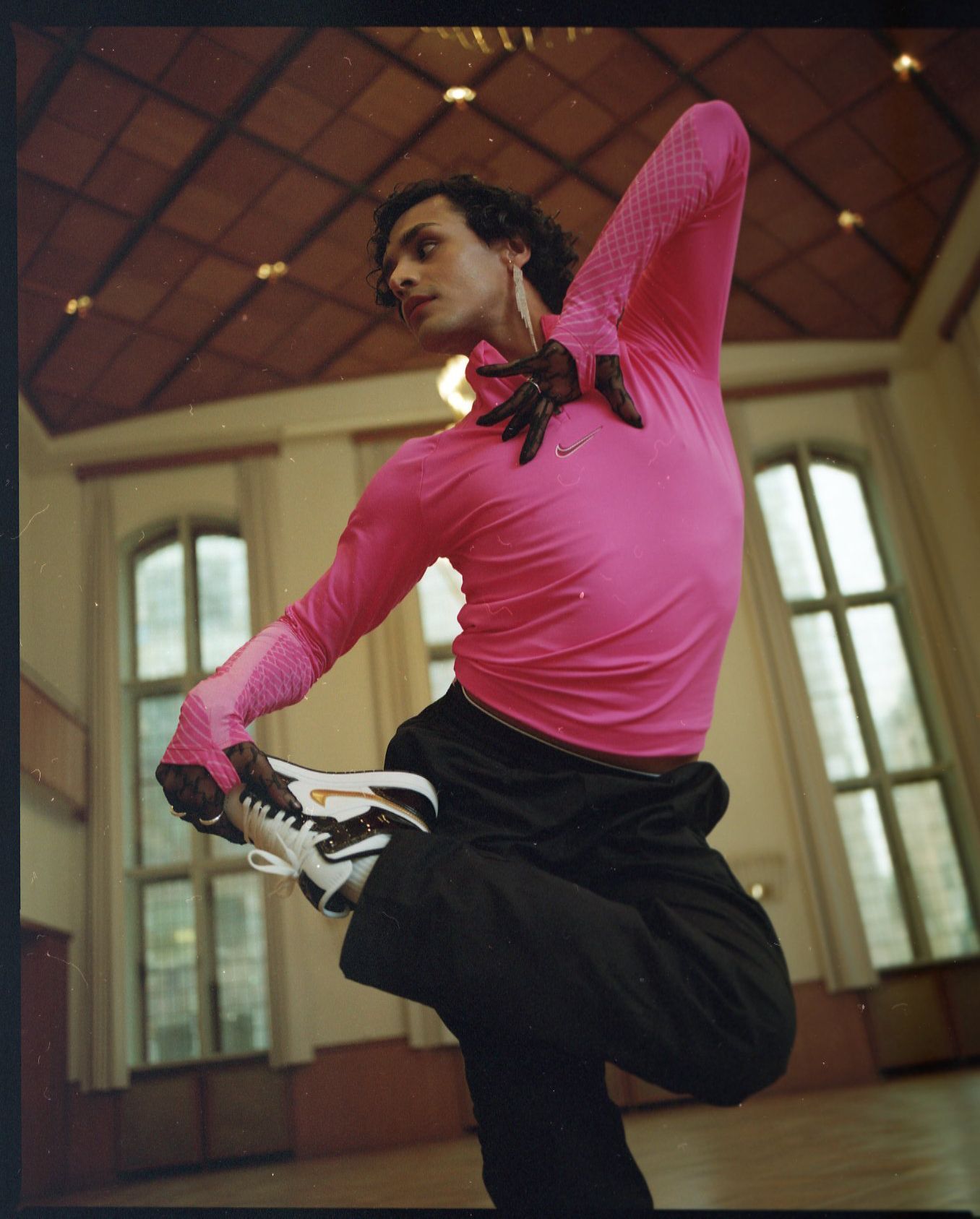Man in pink top and black pants doing a dance pose, wearing sneakers, indoors.