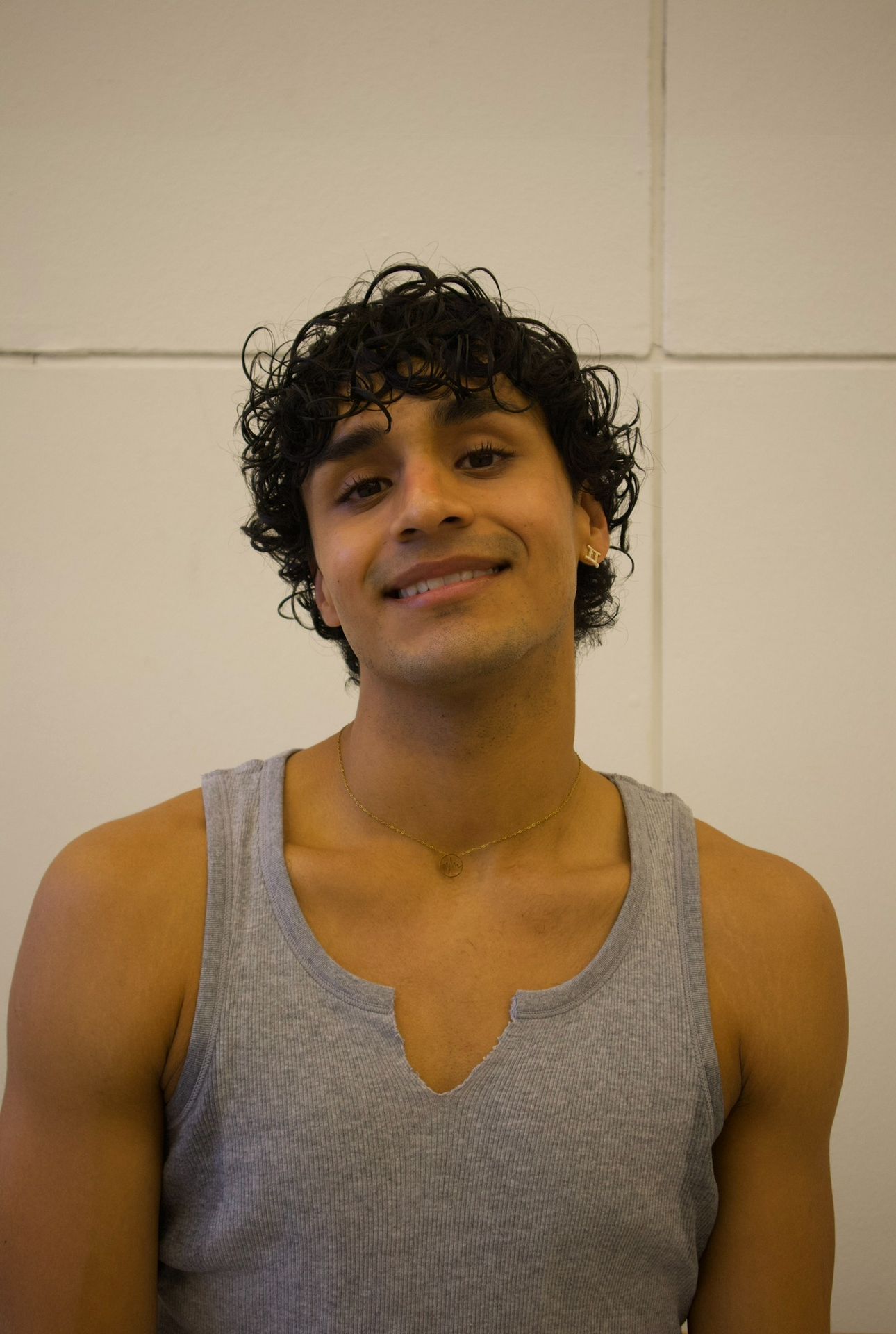 Man with curly hair smiles, wearing a grey tank top against a white background.