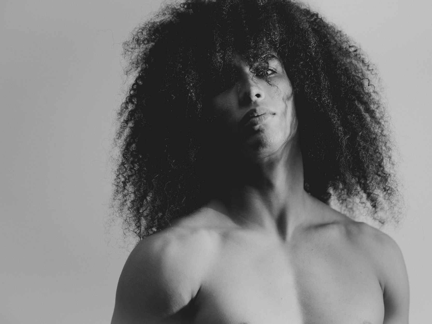 Black and white photo of a person with curly hair, partially shirtless, looking towards the camera.