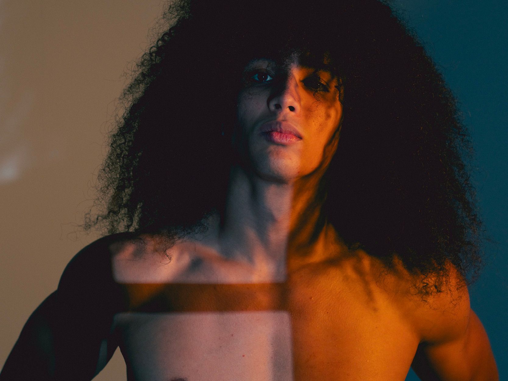 Man with long, dark hair, lit with orange and blue light, gazing forward, torso partially bare.