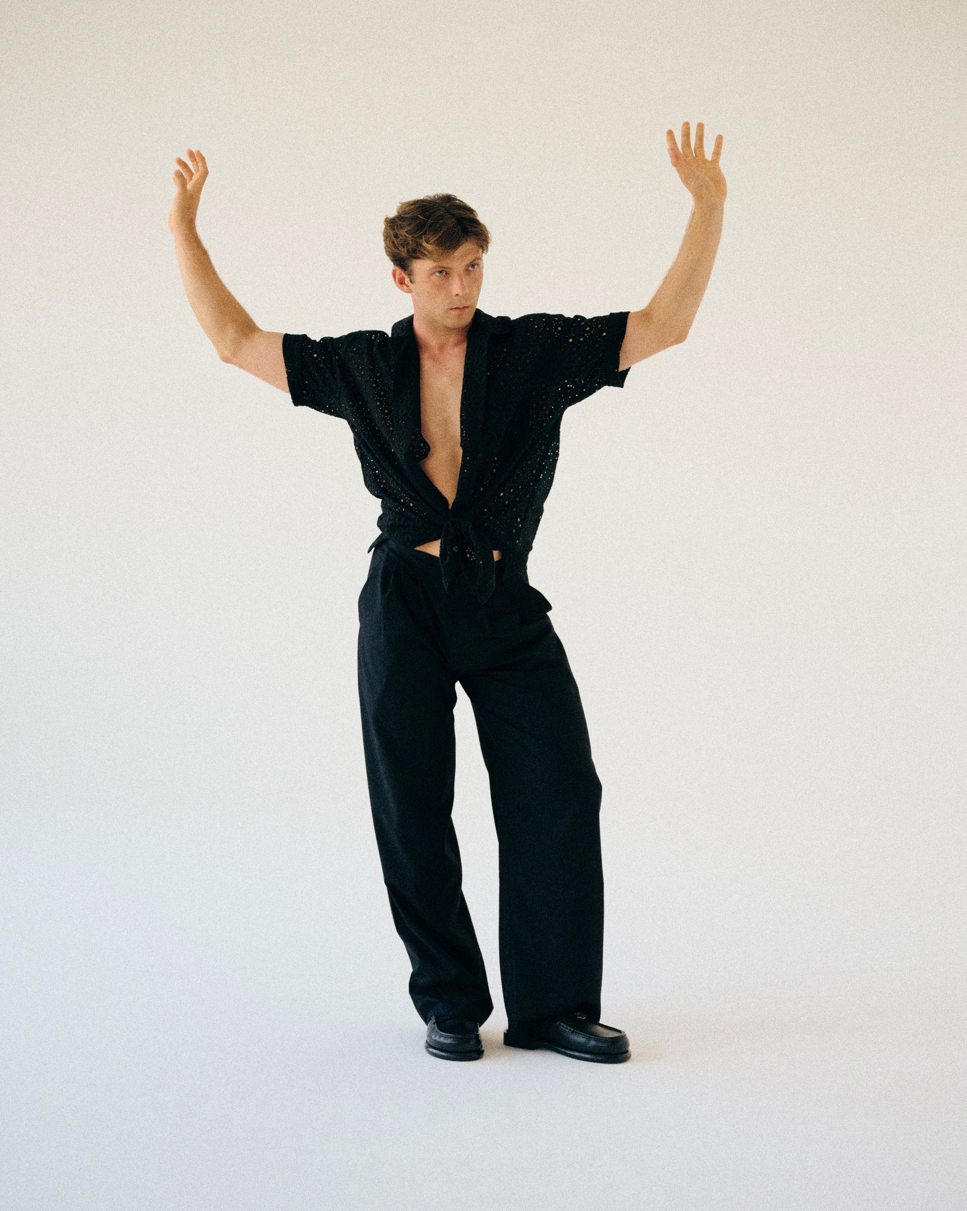 Man in black outfit poses with arms raised against a white backdrop.