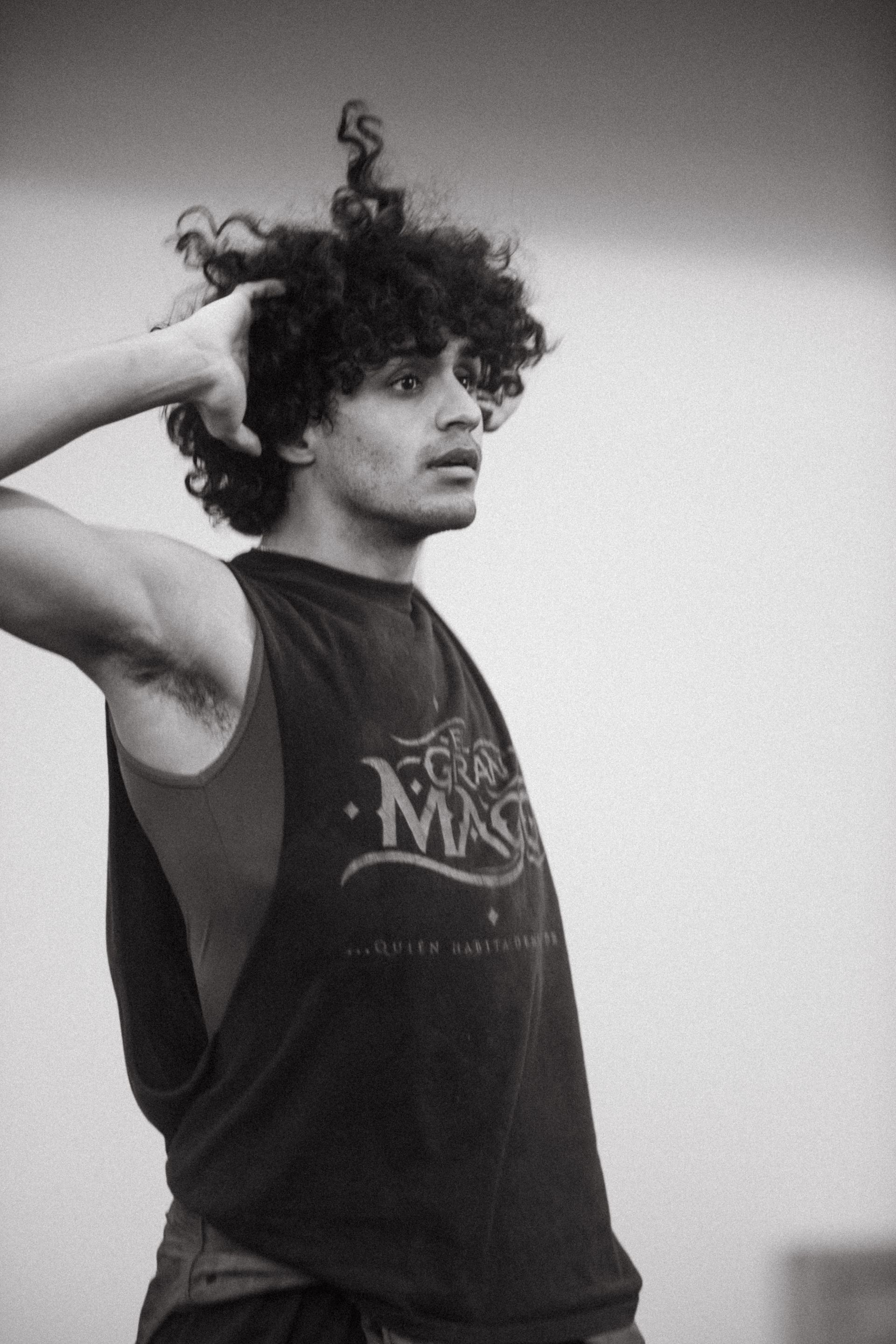 Man in a dark tank top with curly hair touches his head, looking off-screen.