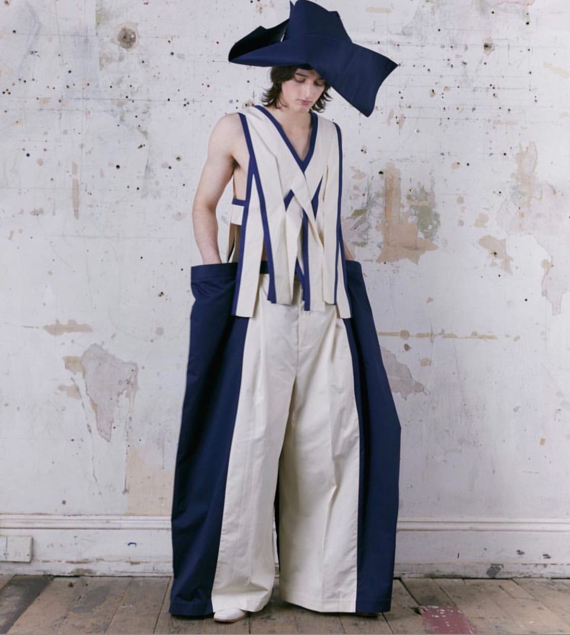 Man in oversized, avant-garde outfit. Navy and cream colors. Large hat, wide-leg pants. Leaning against a distressed wall.