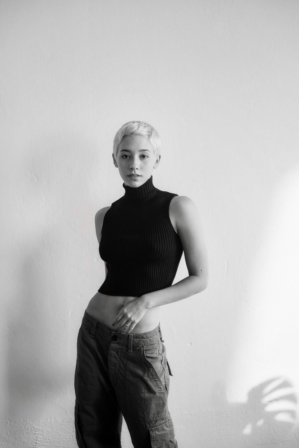 Woman with short blonde hair in black top and cargo pants, standing against a white wall.