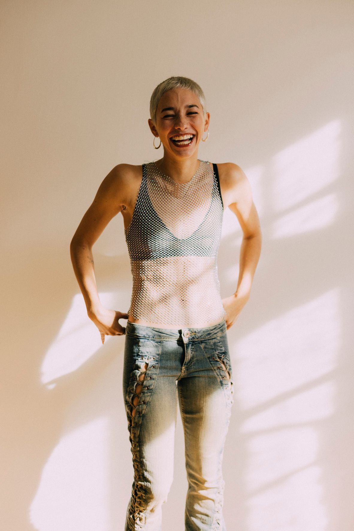 Woman in mesh top and jeans, laughing, hands on hips. White wall with sunlight shadows.