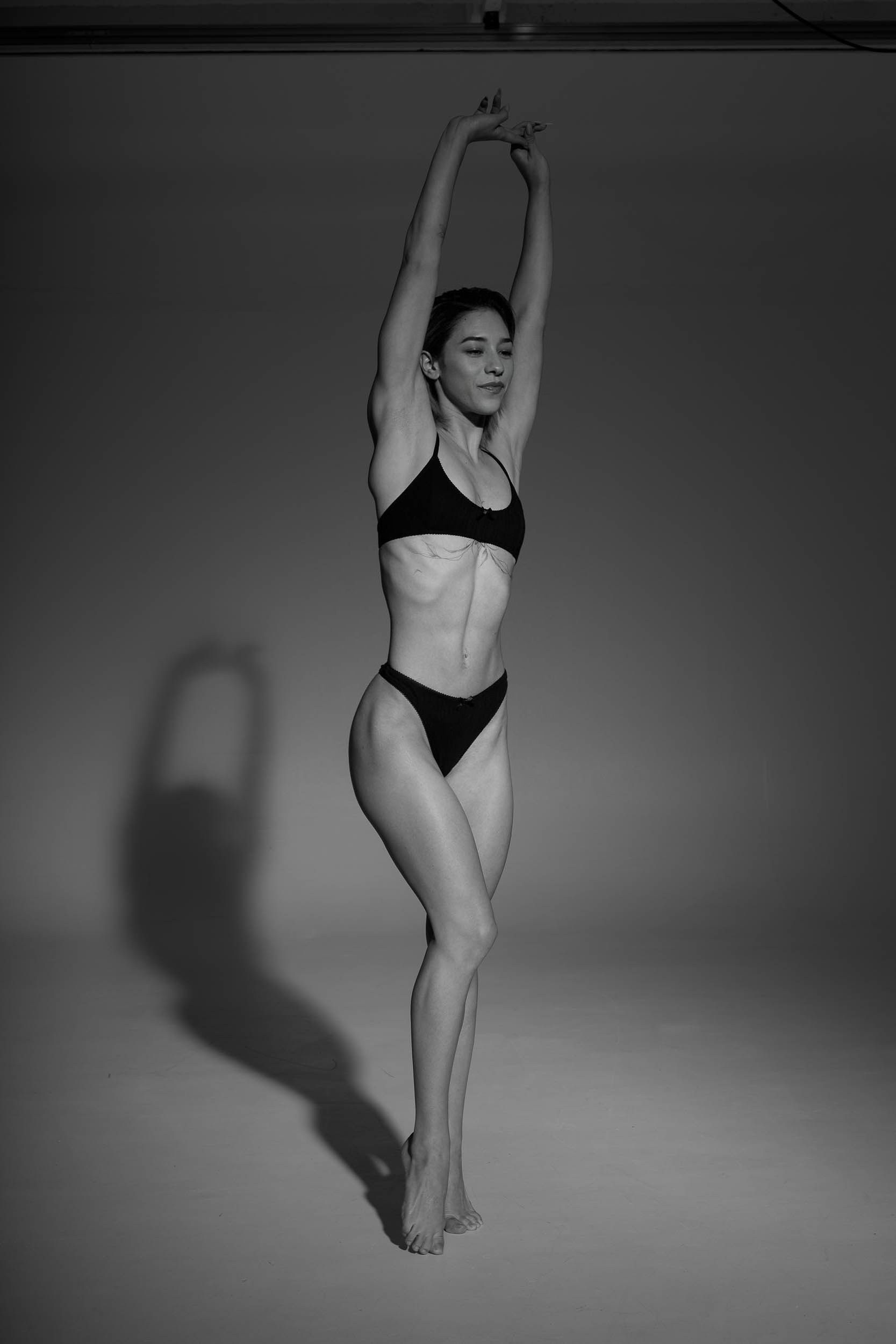 Woman in black lingerie stretches arms overhead in a studio with a shadow.