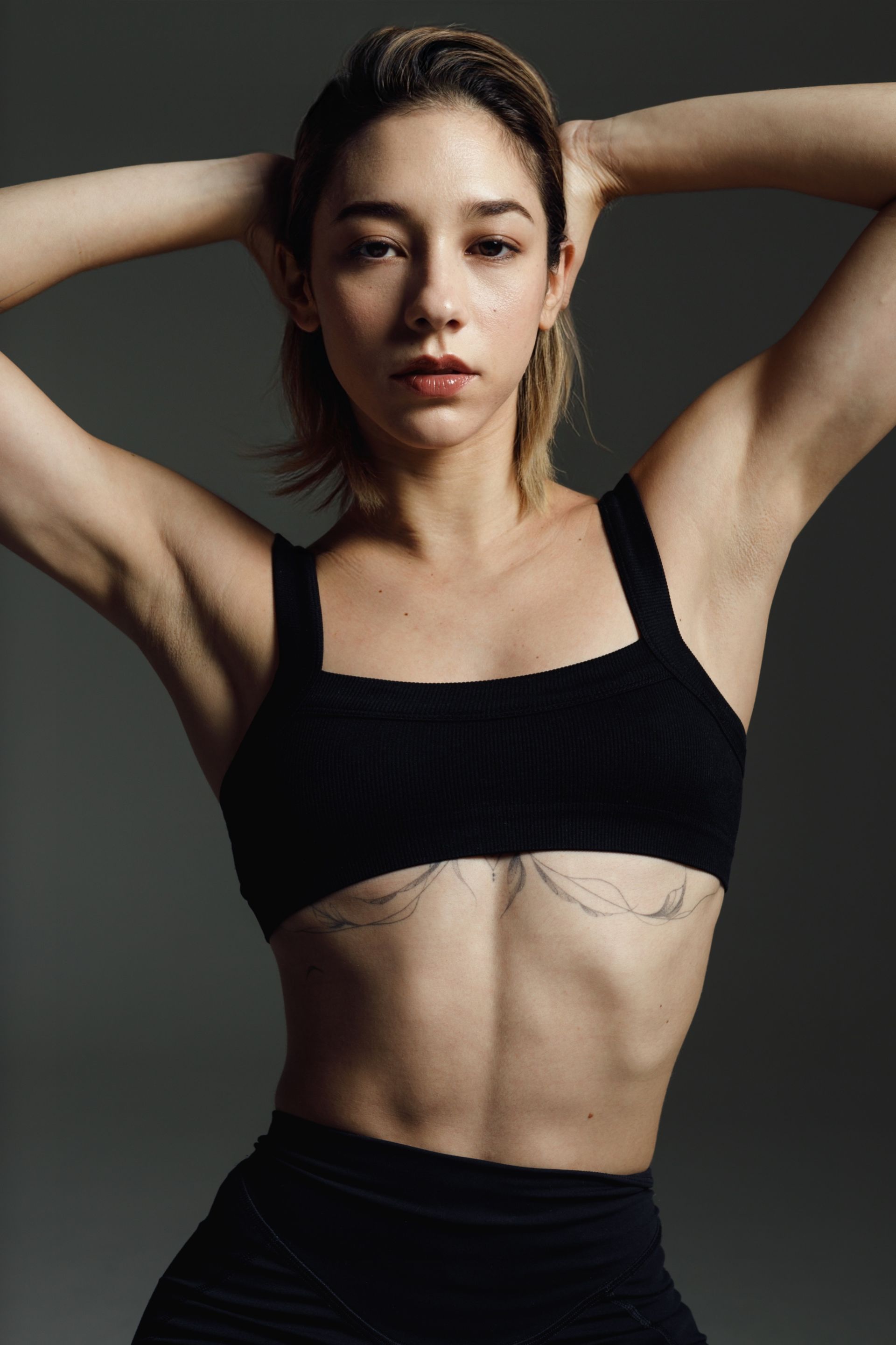 Woman in black sports bra and leggings, hands behind head, looking forward.