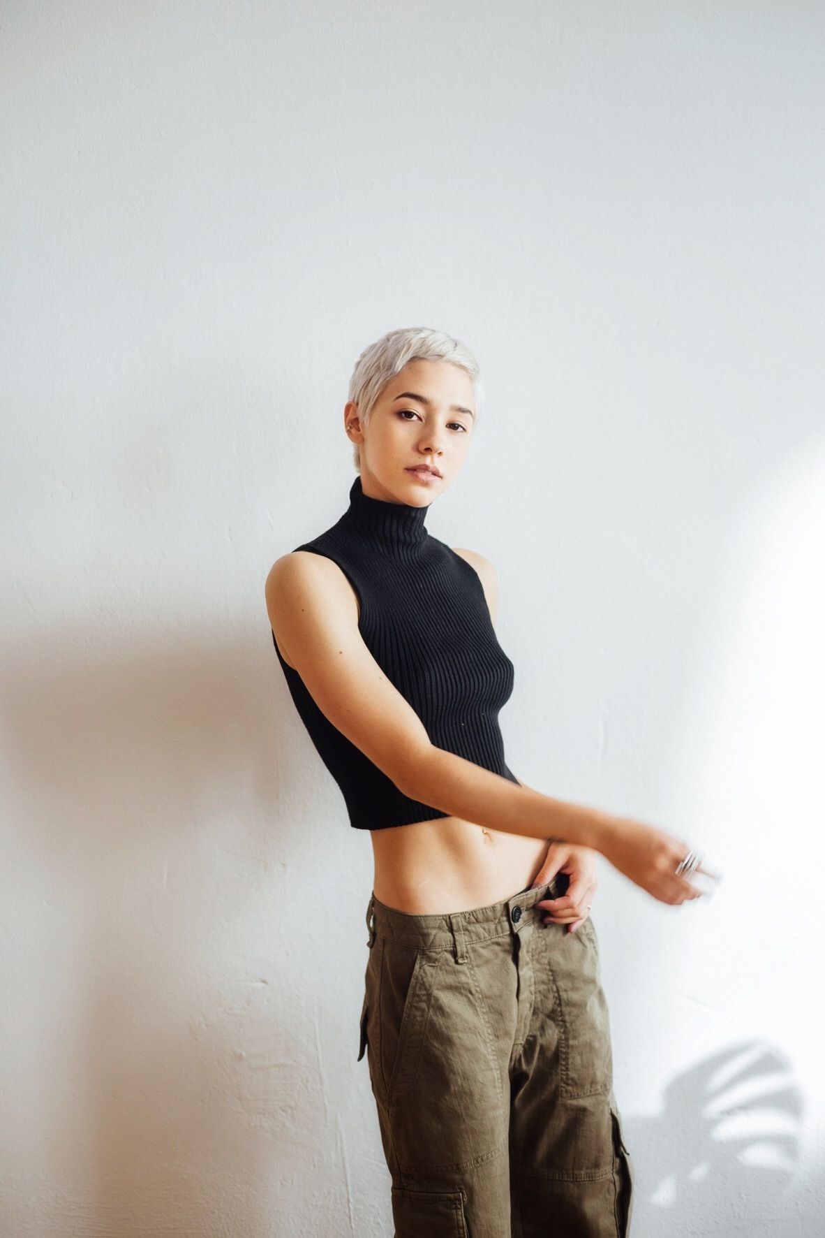Woman with short, light hair wearing a black sleeveless turtleneck and olive-green pants, standing against a white wall.