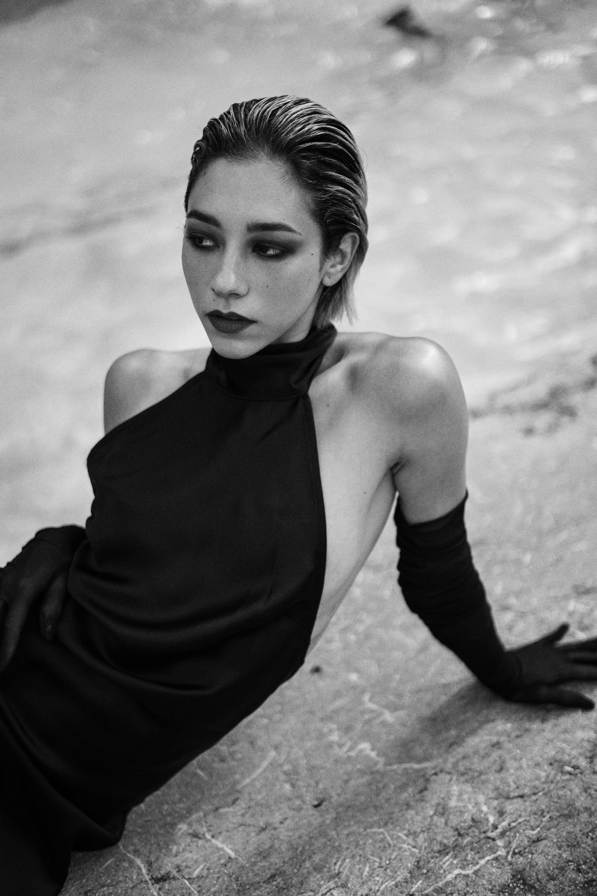 Woman in black dress and gloves, posing by water.