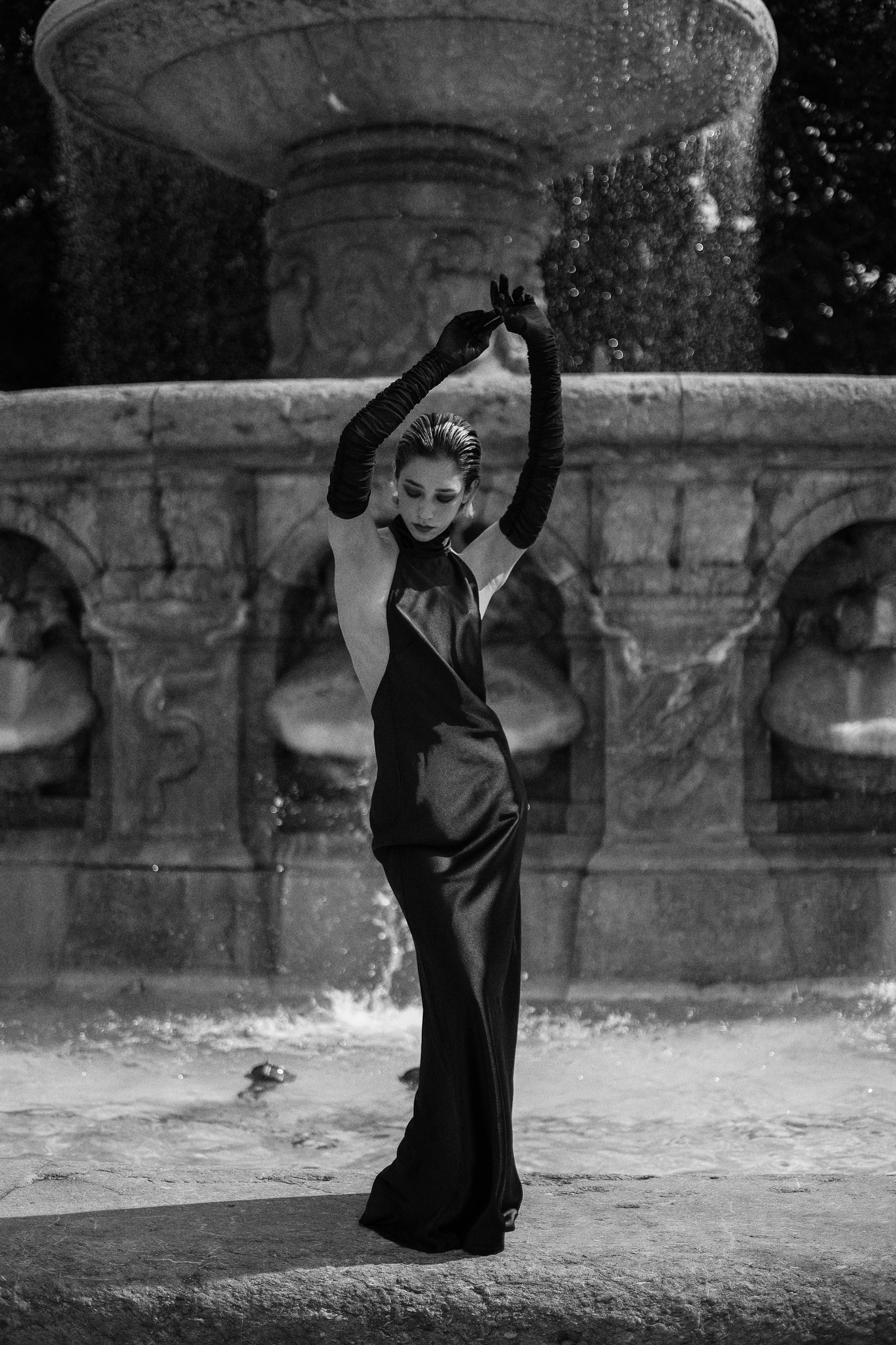Woman in long black dress and gloves poses dramatically by a fountain.
