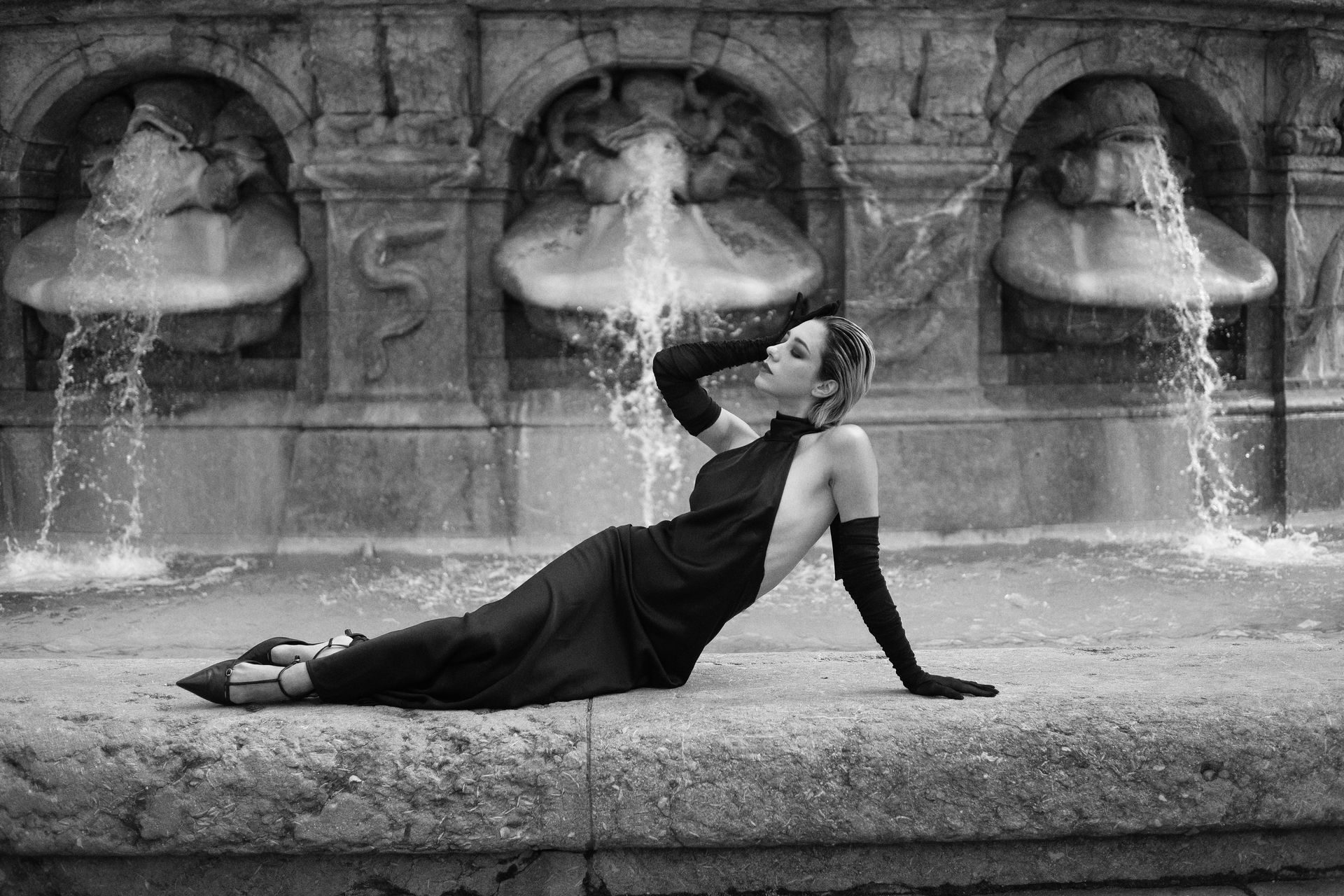 Woman in black dress and gloves reclines near ornate fountain, arms raised, water flowing.