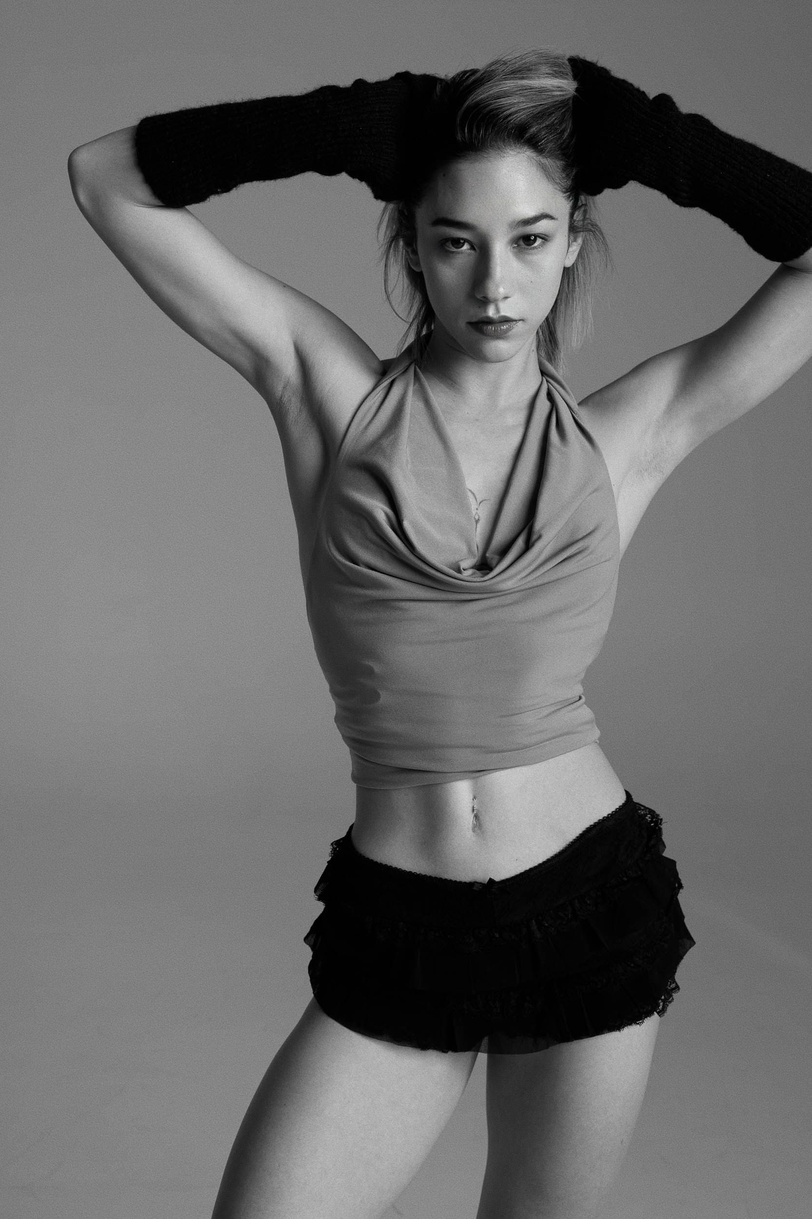 Woman in halter top and shorts, hands above head, looking at the camera. Black gloves. Studio shot.