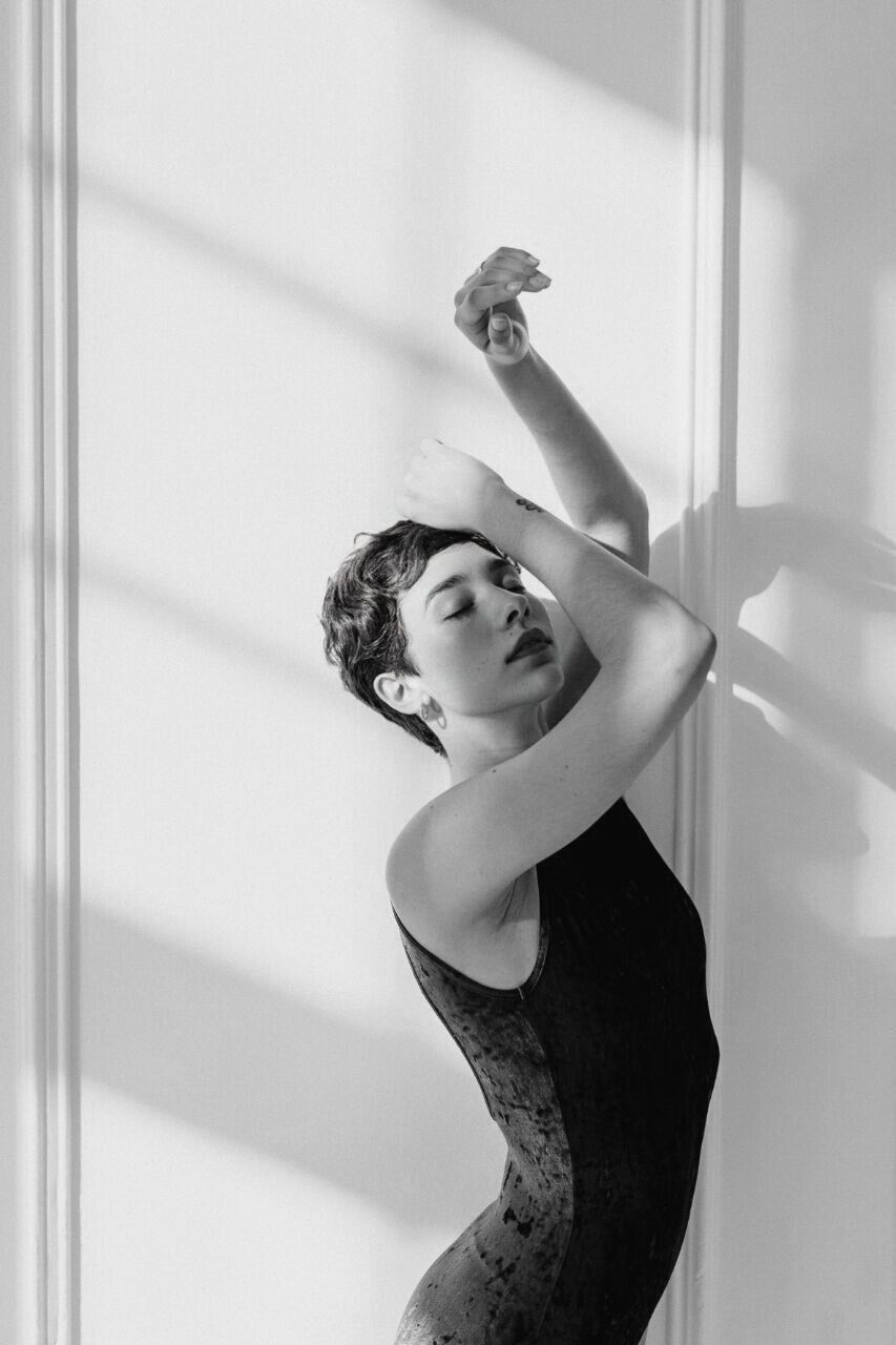 Woman in black leotard leaning against a white wall, arms raised, eyes closed, sunlight creating shadows.