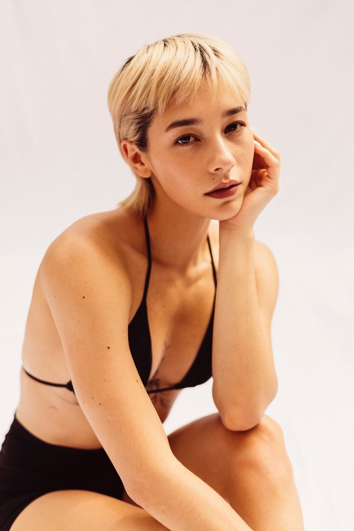 Blonde-haired person in a black bikini top and shorts, leaning on hand, looking at the camera.